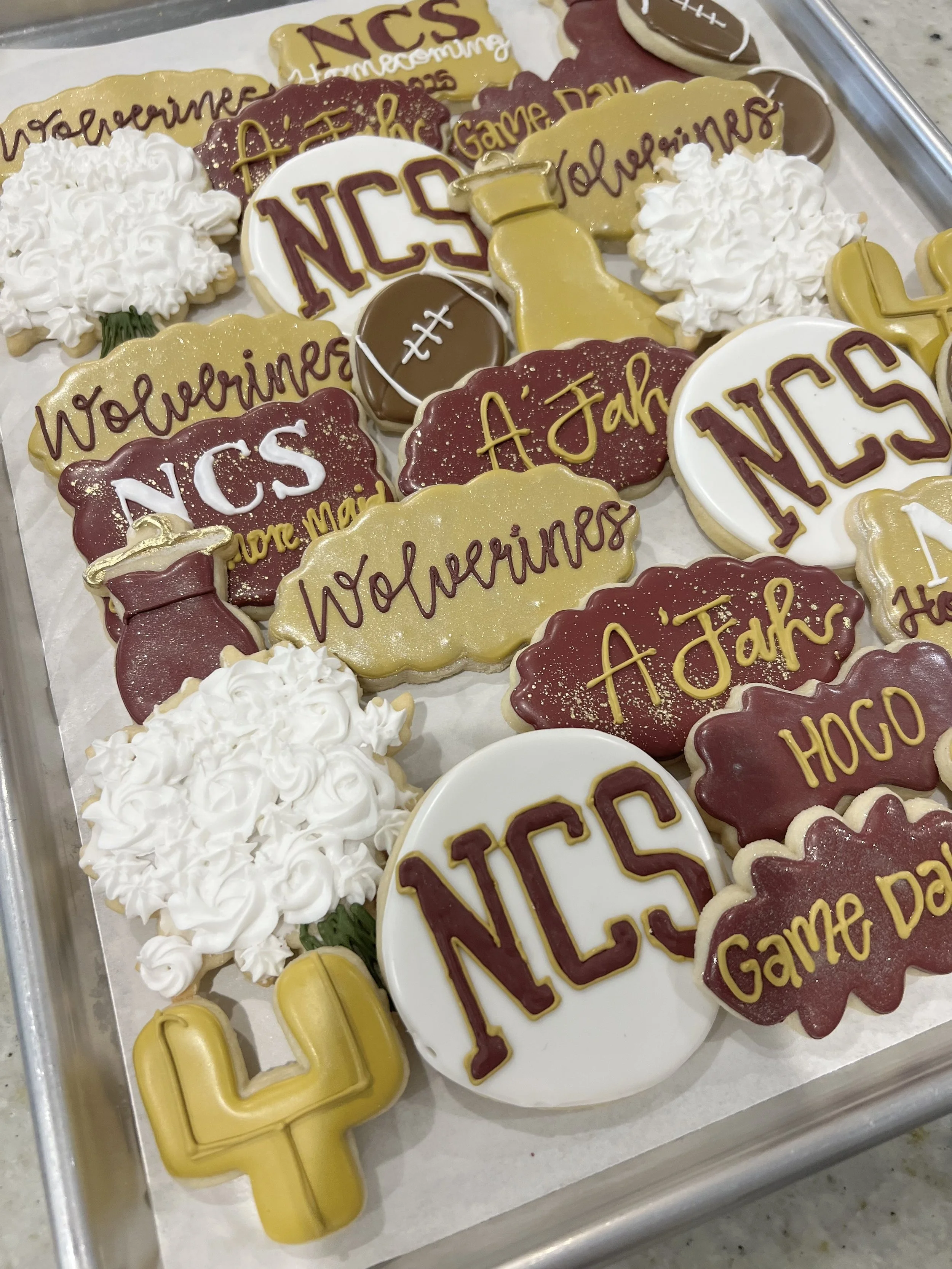 Assorted decorated cookies celebrating a football game day, featuring items like cookies with the NCS logo, football shapes, pennants, cheerleaders, with messages like "A Job" and "Game Day."