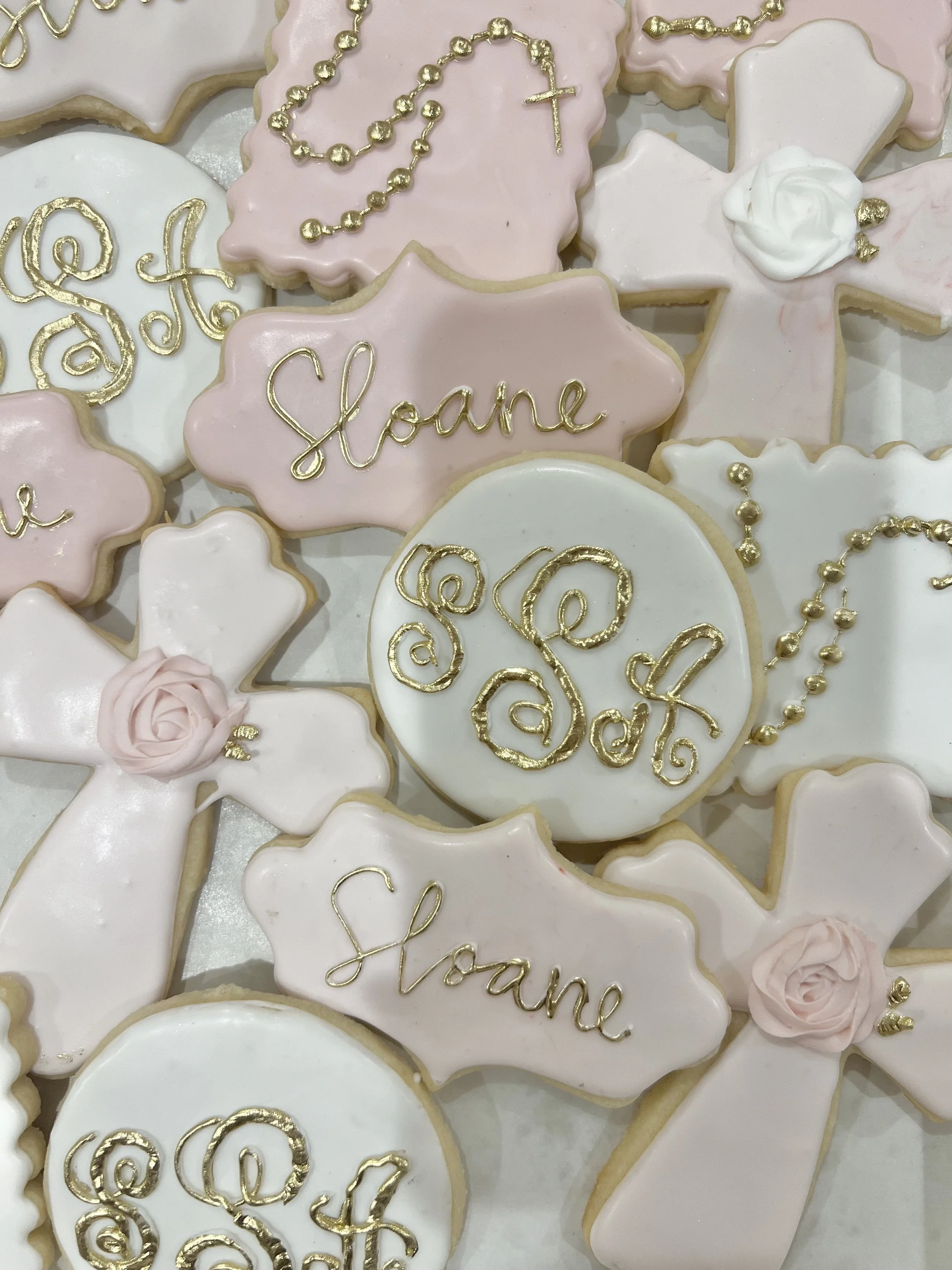 Decorated cookies with pink and white icing, shaped as crosses and ovals, with gold lettering and embellishments, some with pink roses and chains.