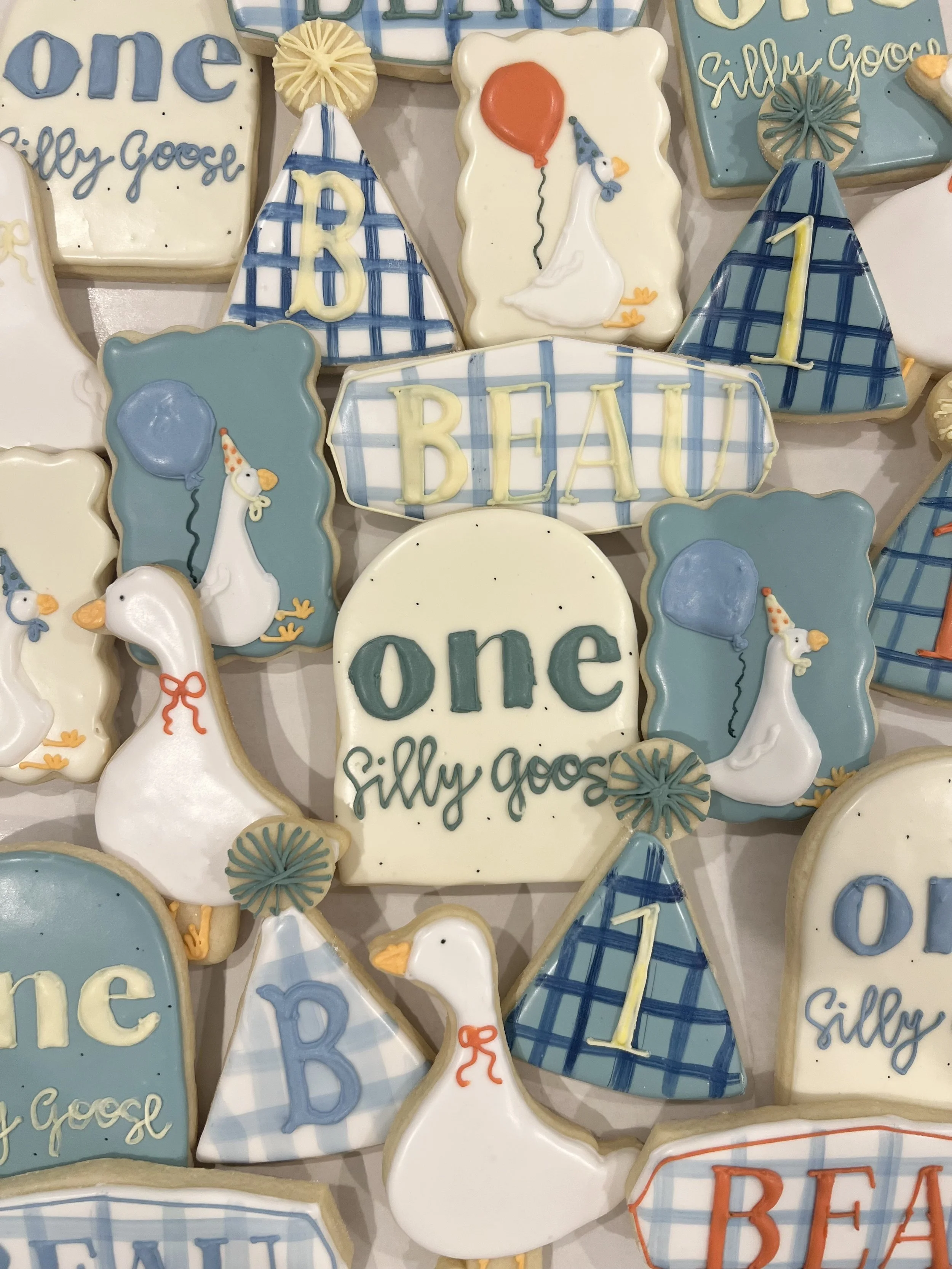 Decorative cookies with birthday themes, featuring swans, balloons, and the words "one," "BEAU," "B," and "silly goose" in pastel colors and various patterns.
