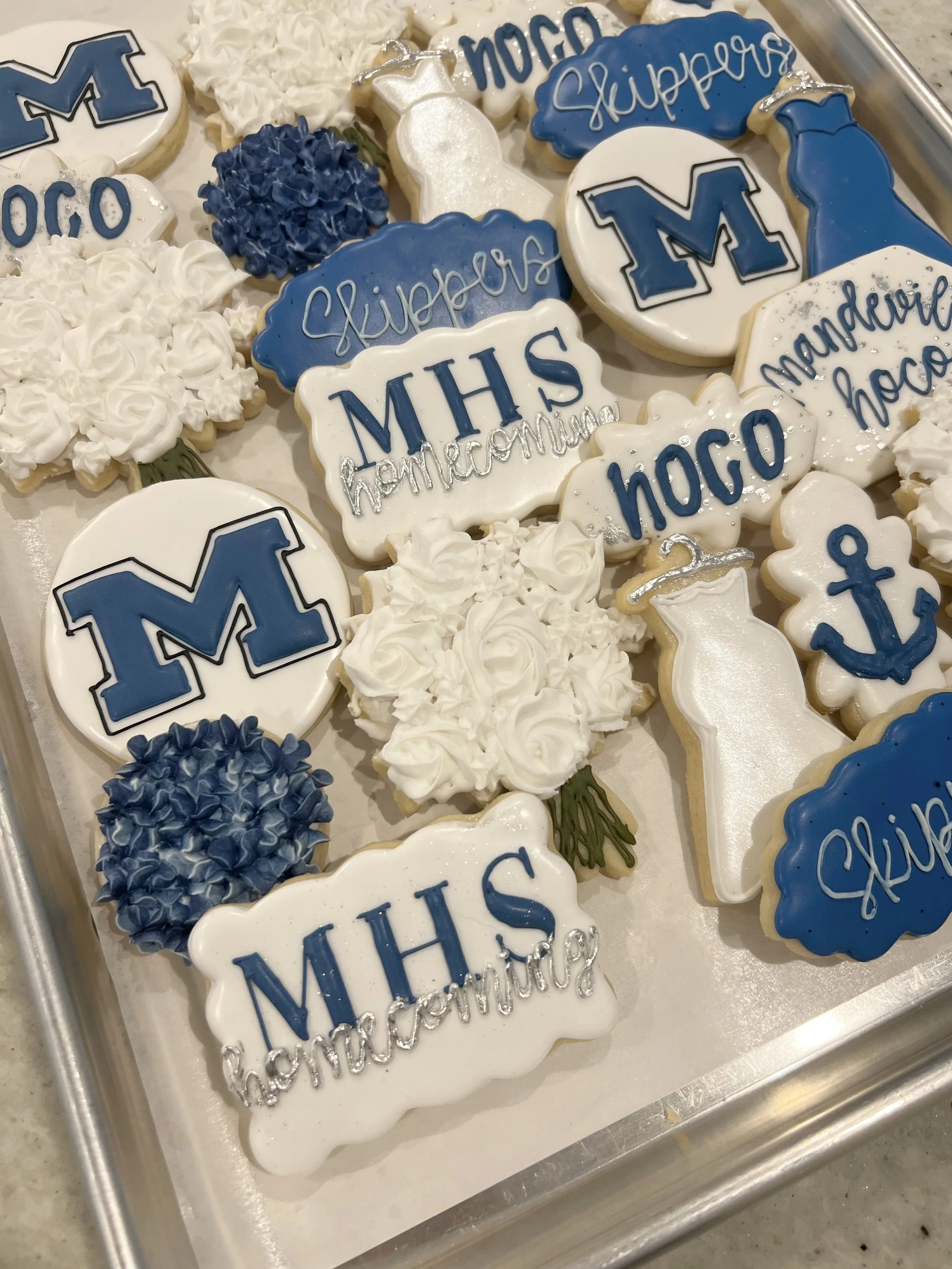 Mandeville High School collection of decorated cookies with blue, white, and silver themes, featuring words like "skippers," "MHS homecoming," and "hoco," along with nautical symbols such as anchors, and floral designs.
