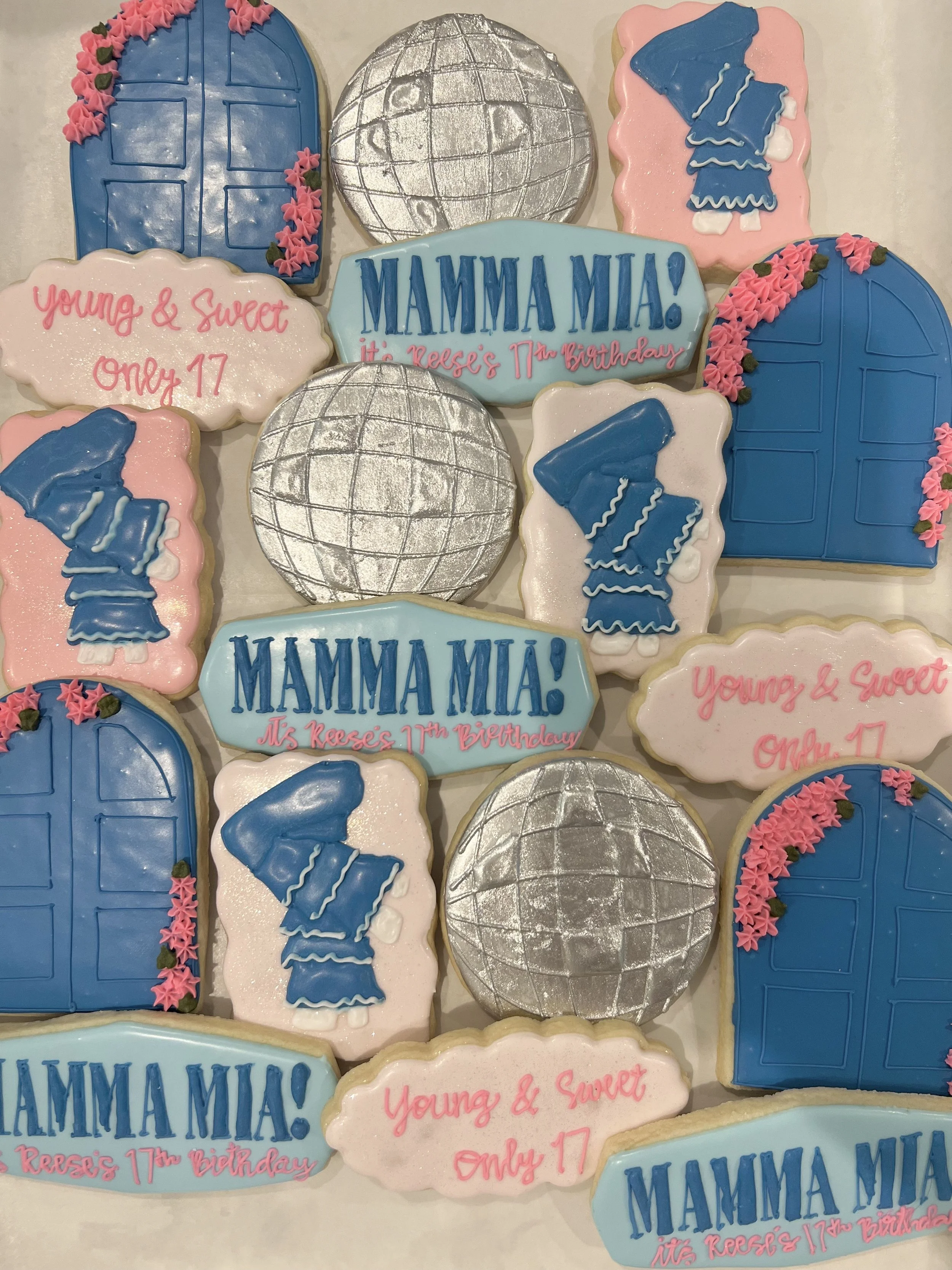 Cookies decorated with themes from the movie 'Mama Mia' and a 17th birthday celebration. The cookies include pink and blue dresses, disco balls, door decorations, and banners with graduations and birthday messages.