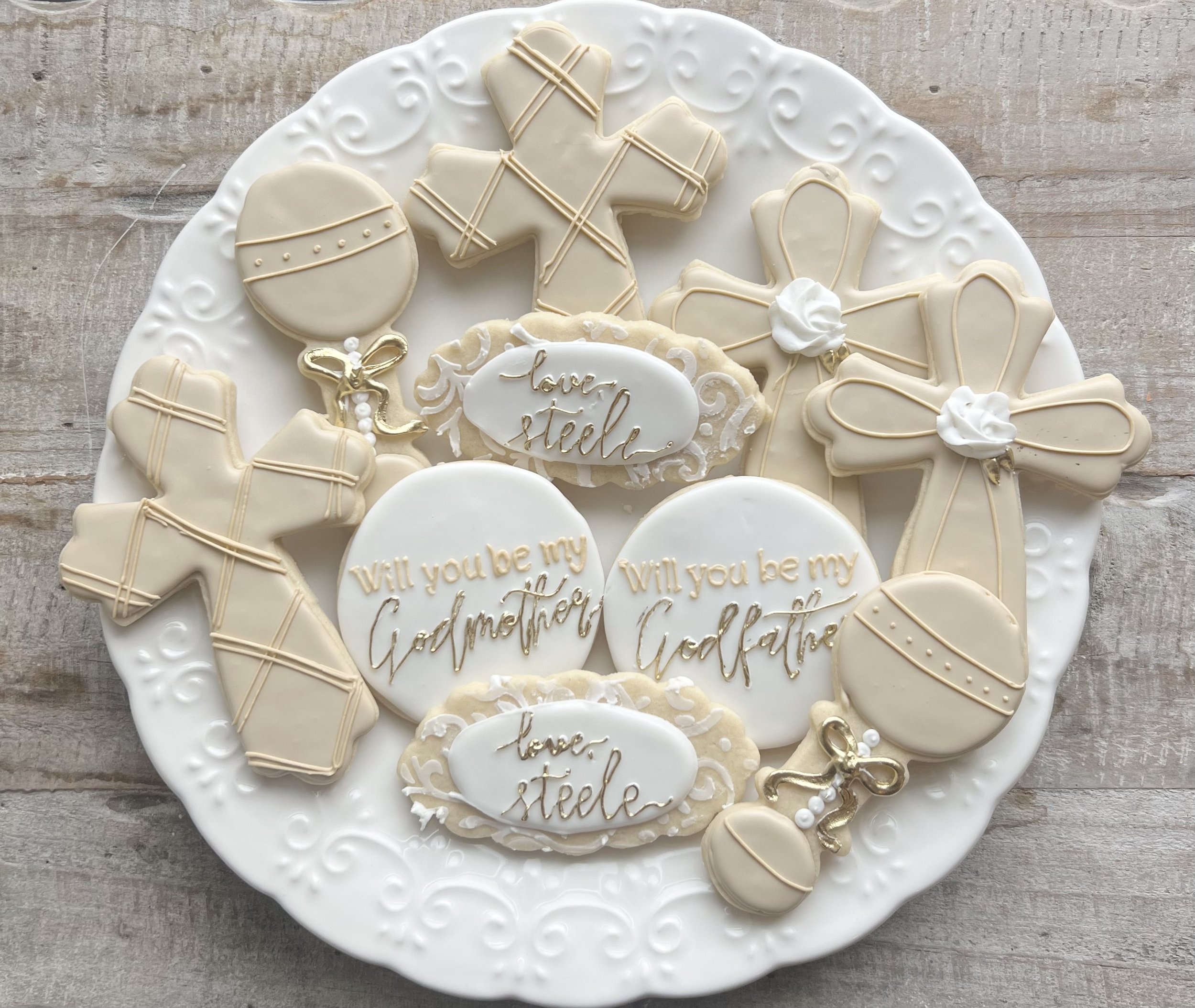 Decorated sugar cookies on a white plate with floral and swirl designs, featuring messages like 'Will you be my Godmother' and 'Love, Stella' in gold writing, along with gingerbread men, bows, ornaments, and a small gold baby rattle.
