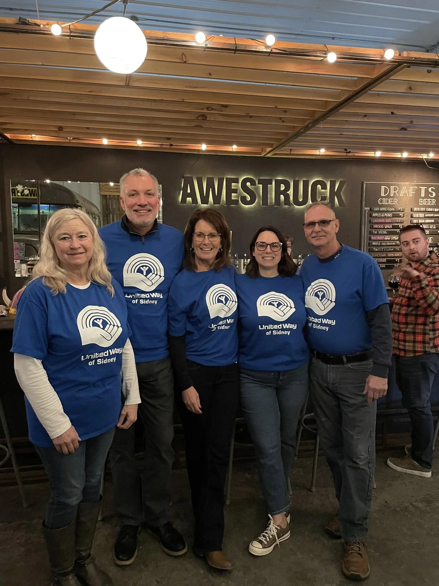 Group of six people standing together indoors, all wearing blue United Way of Sidney t-shirts, smiling for the photo in front of a dark wall with a neon sign that reads 'AWESTRUCK'. One person in the background is holding a glass of wine.