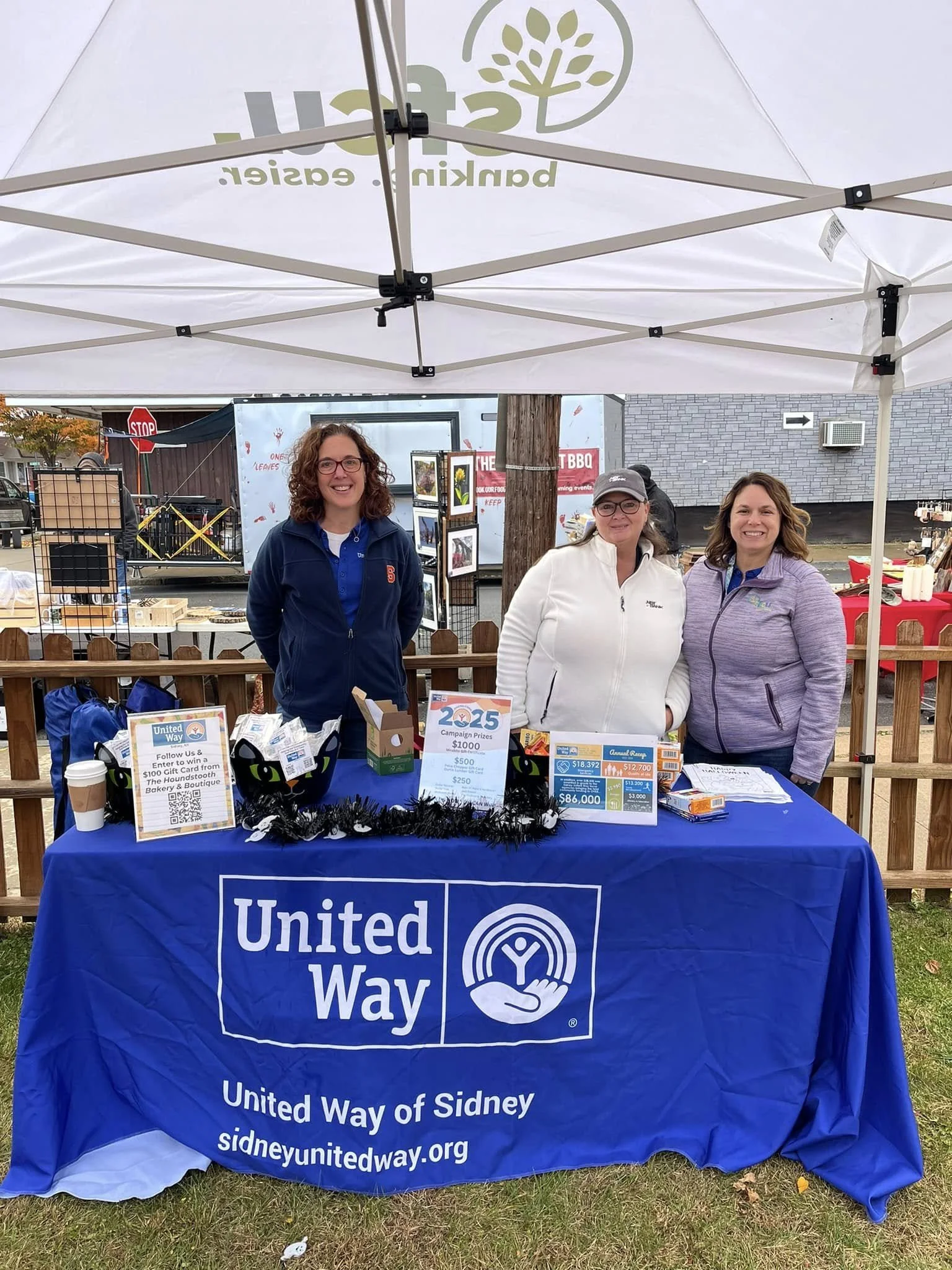 Three women standing behind a United Way table outdoors at a fundraising event, smiling, under a white canopy, with promotional materials and donation signs on the table.