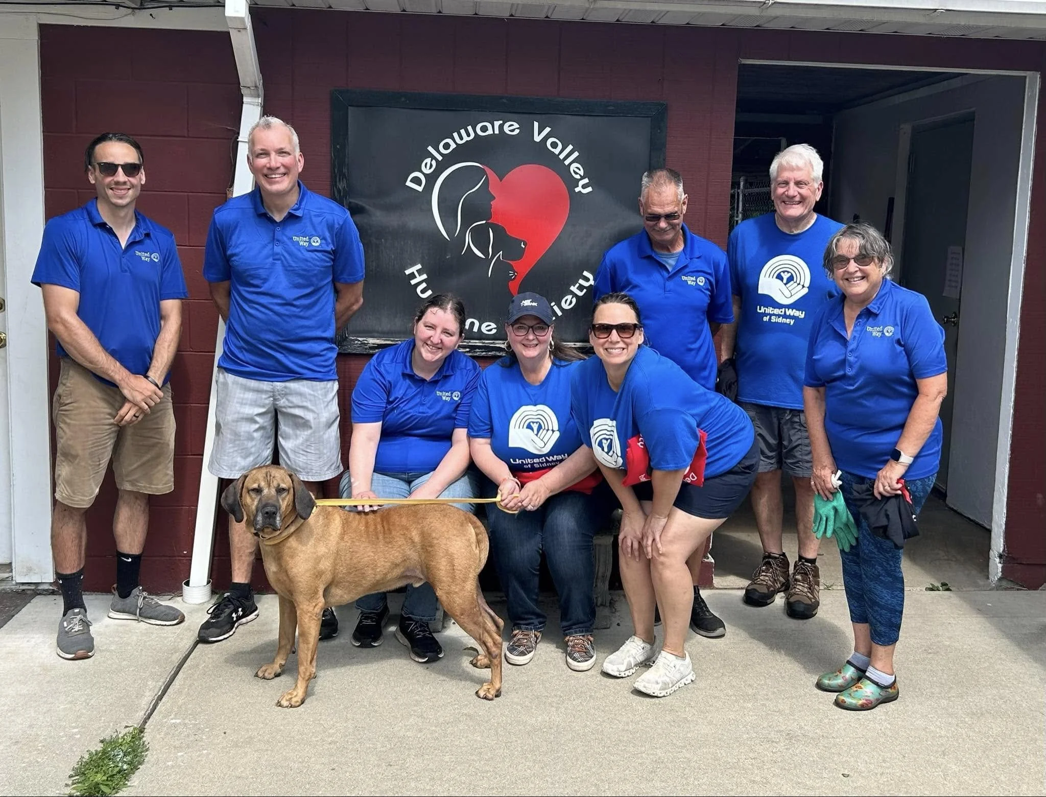 Group of nine people, including a dog, standing outside in front of a red building with a heart and dog silhouette logo for Delaware Valley Humane Society. All are wearing blue shirts, some with United Way logos, smiling for the photo. The dog is light brown and standing on leash.