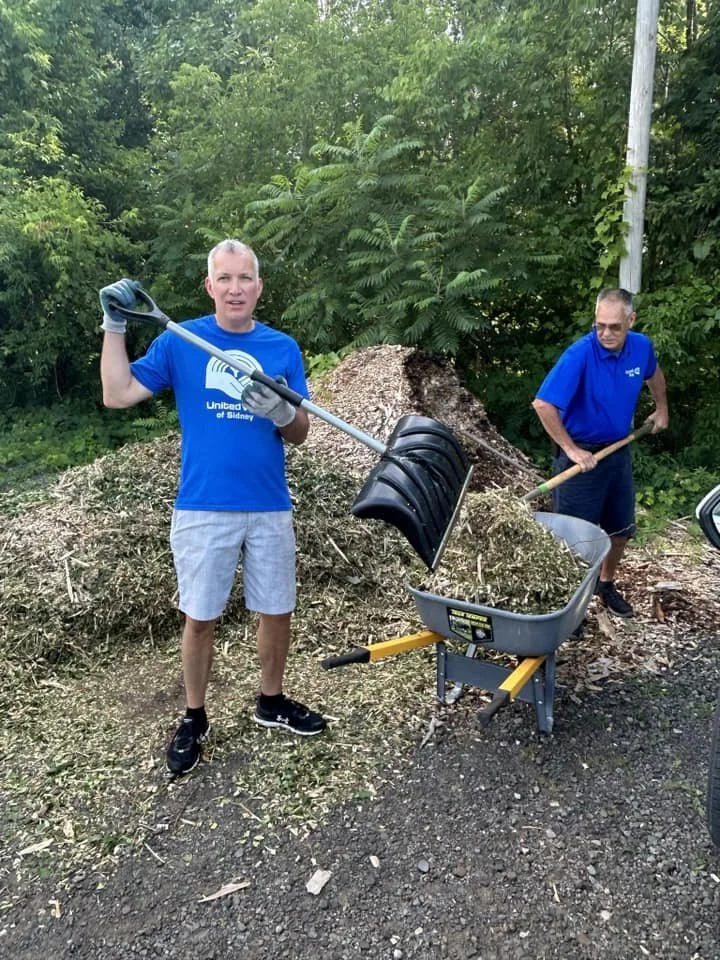 Two men working outdoors near a large pile of wood chips or mulch, using tools. One man holds a shovel, and the other manual tool, with greenery in the background.