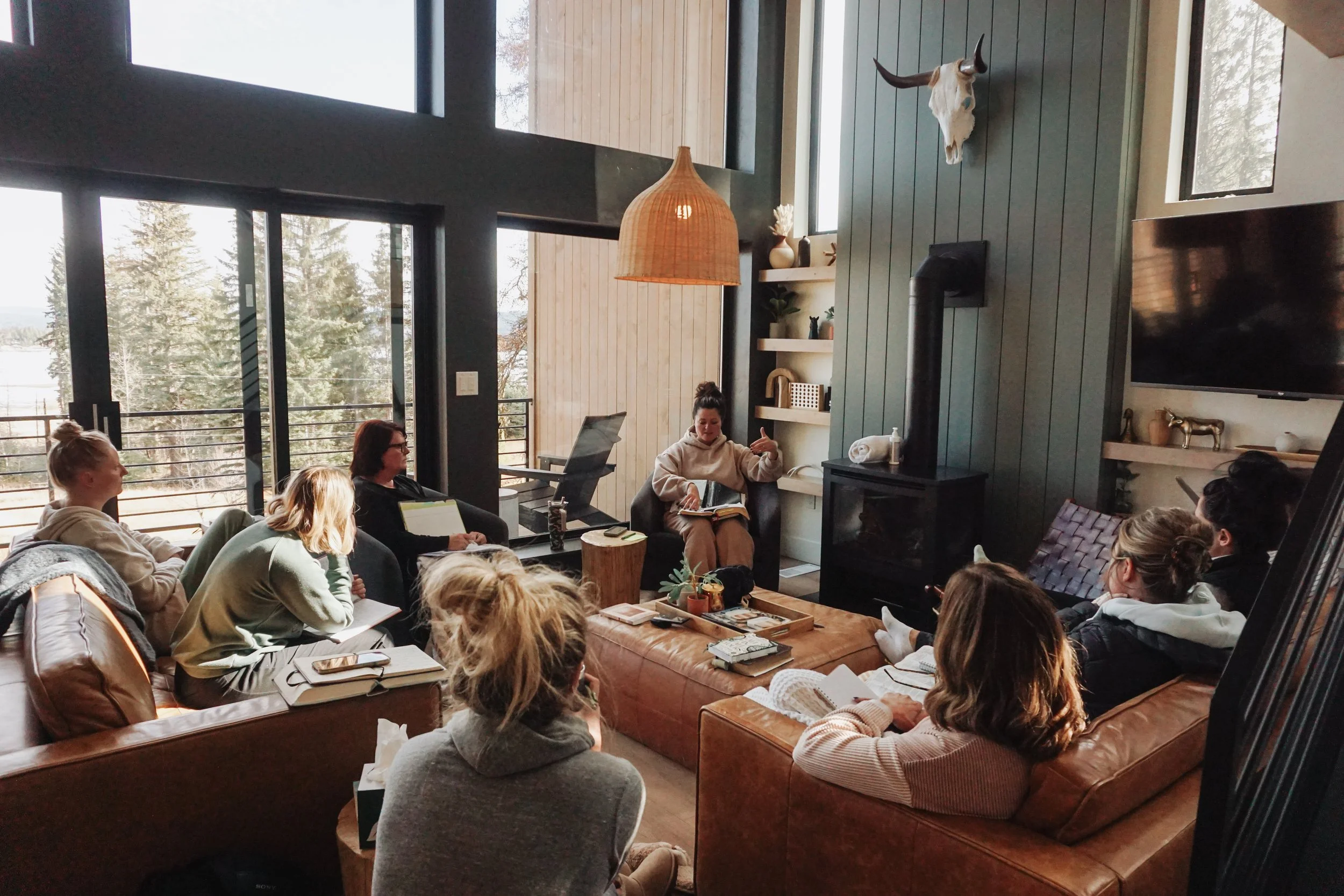 A group of women sitting in a cozy living room, engaged in a discussion with a woman who appears to be leading the meeting; the room has large windows, a wood stove, and decorative items.