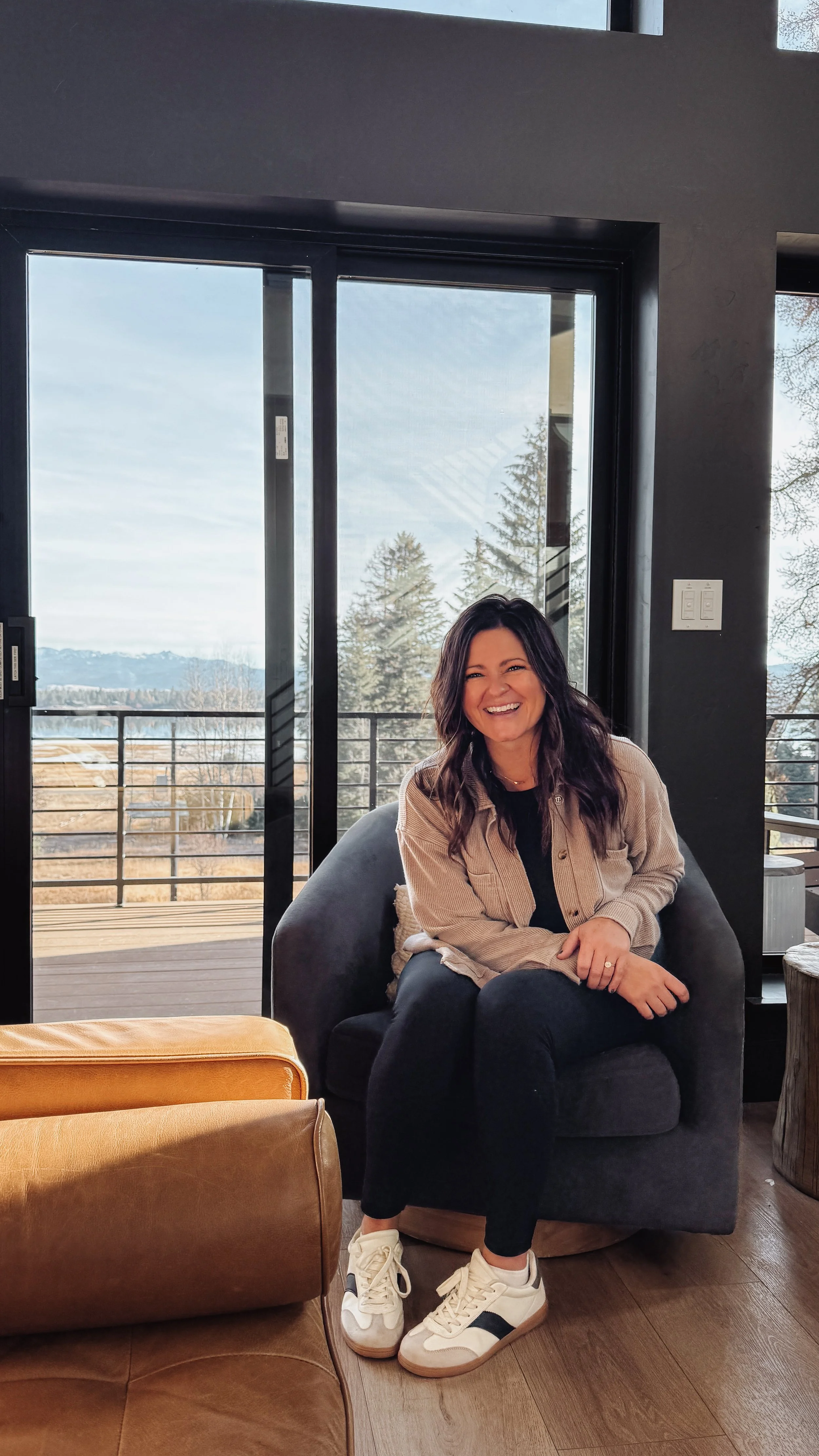 A woman sitting on a black armchair near a large glass door with a scenic outdoor view of trees and mountains, smiling and wearing casual clothes, including sneakers.