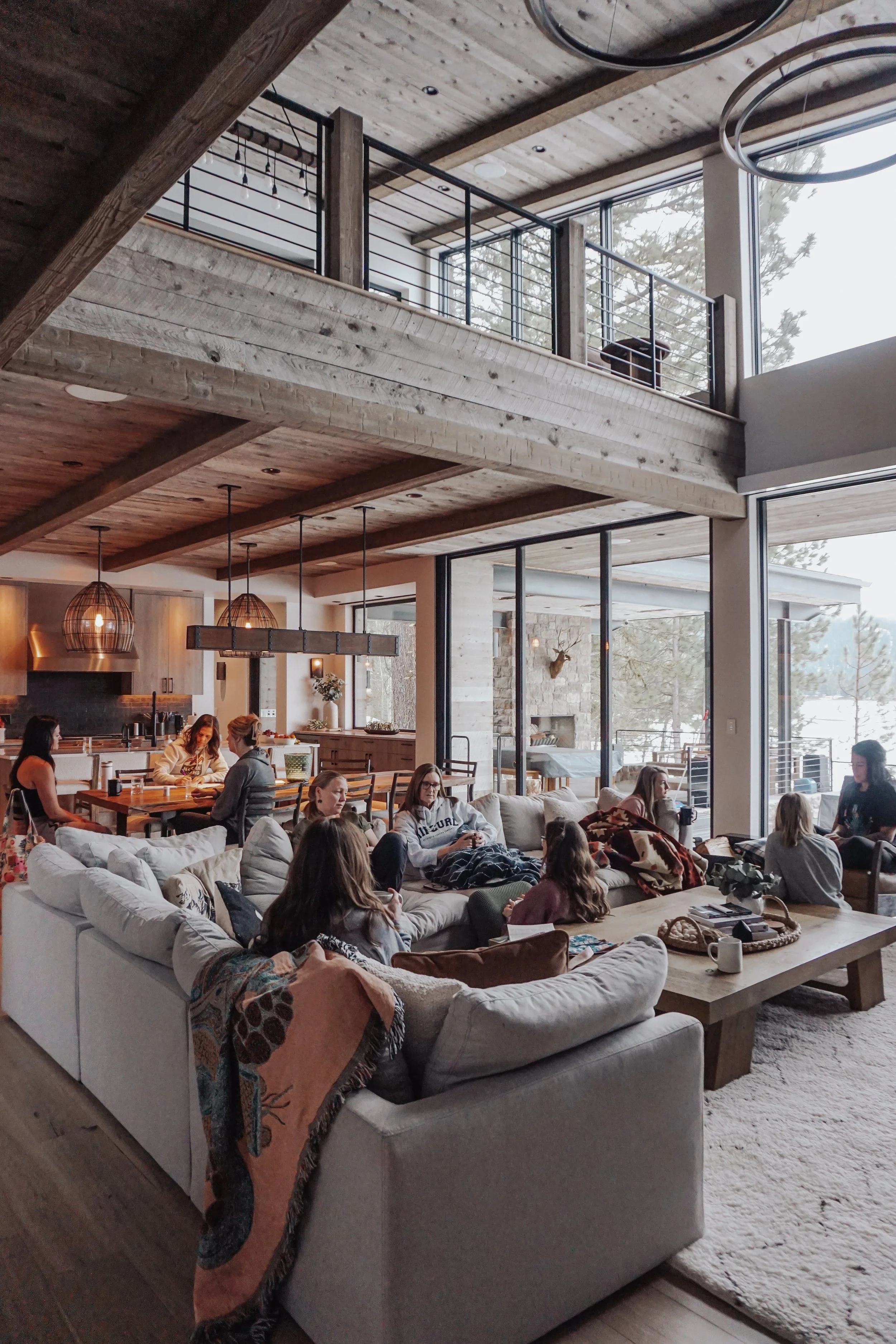 A spacious living room with a large white sectional sofa, wooden coffee table, and large windows overlooking snowy outdoor scenery. Several people are sitting on the sofa and at the dining table, engaging in conversation or using their phones. The room features wood accents, modern lighting fixtures, and a view of the kitchen area in the background.