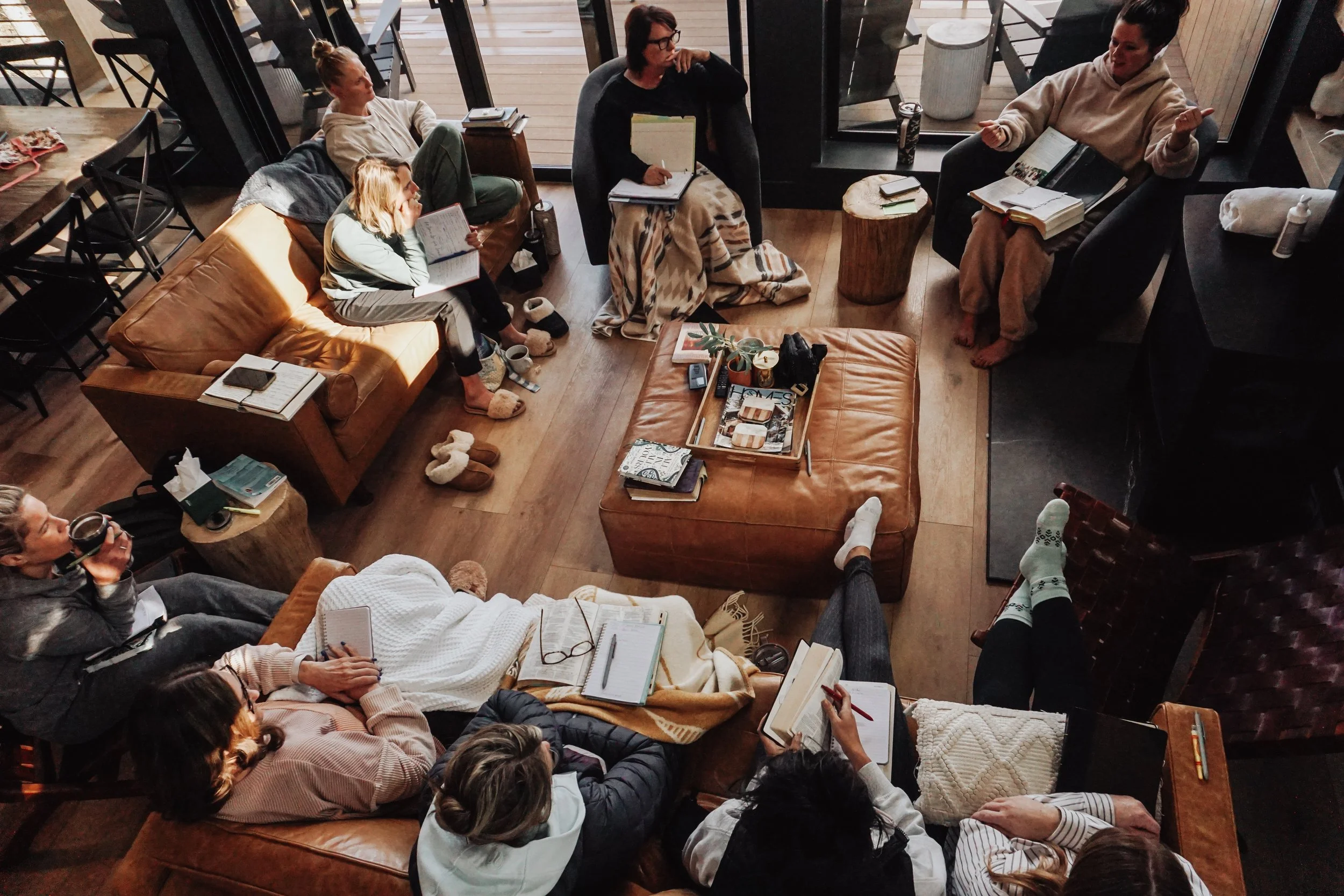Group of people sitting in a cozy living room, engaged in a discussion or reading, with some taking notes. The room has large windows with a view of a wooden deck outside, and various books and cups are visible on tables and cushions.