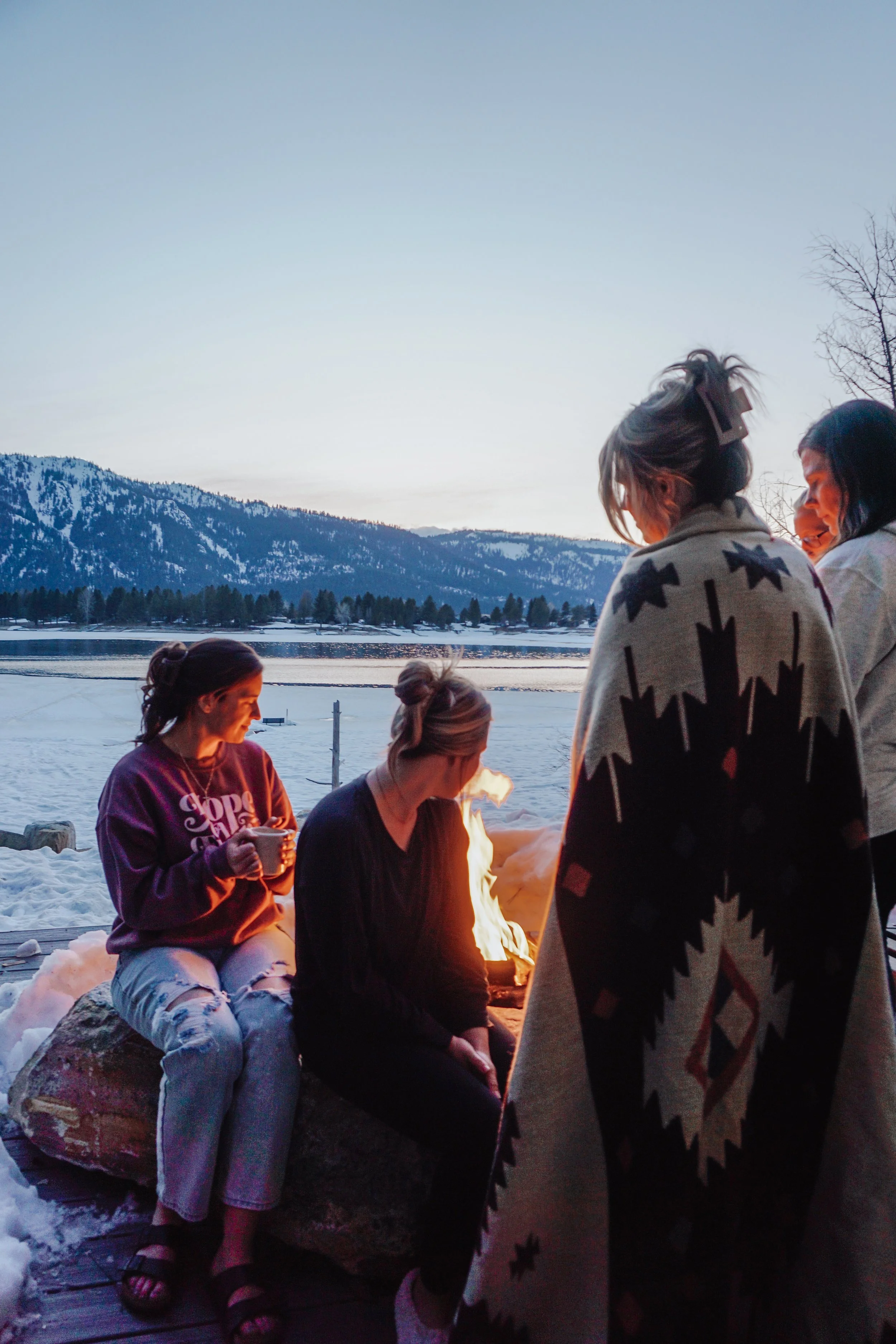 Four women gathered around a fire pit outdoors during winter, with snow on the ground and mountains in the background. The women are dressed warmly, some in sweaters and one in a blanket, enjoying the evening.