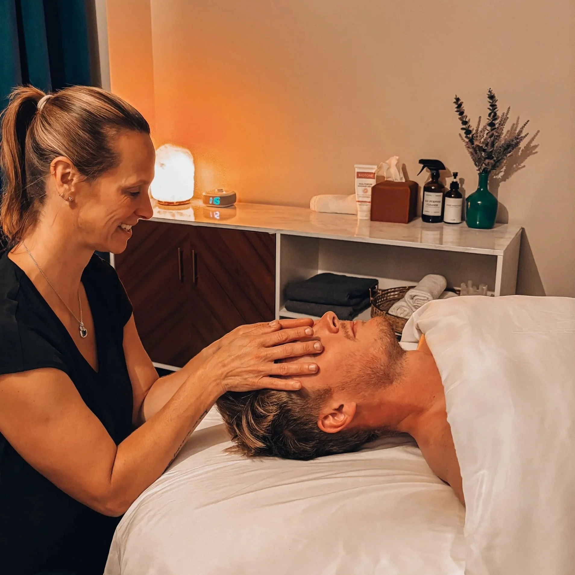 A woman providing massage therapy to a man lying on a bed in a spa or clinic setting, with soothing lighting and spa items visible in the background.