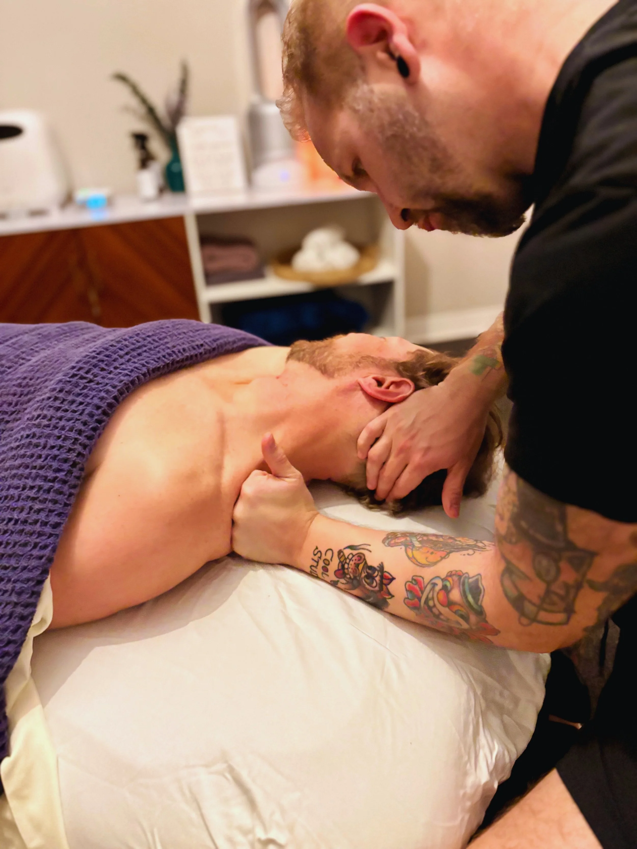 A man with tattoos and earrings performs a massage or chiropractic treatment on a woman lying face up on a massage table, covered with a purple towel.