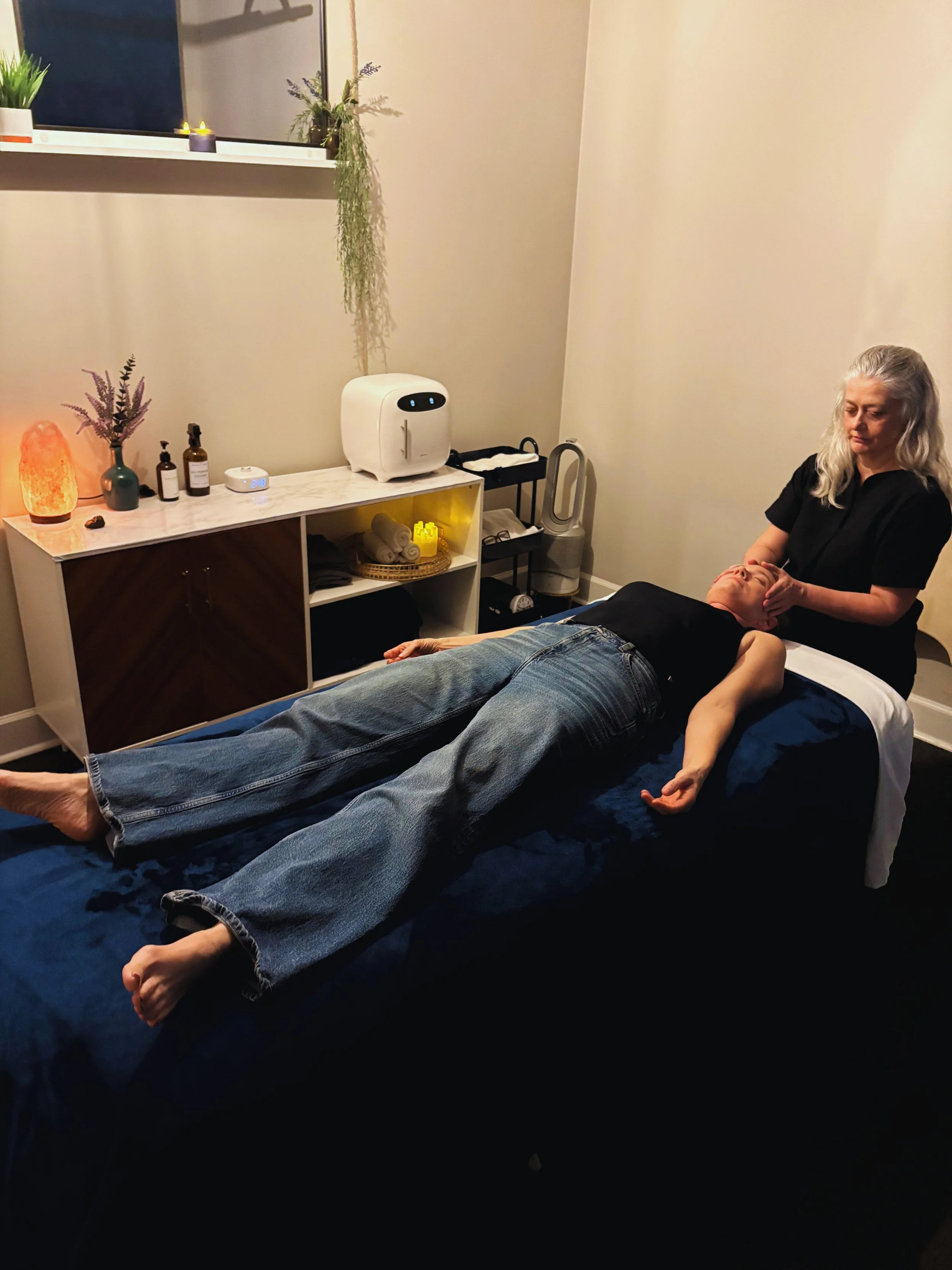 A woman receiving an eye treatment while lying on a massage table in a wellness room with minimalistic decor, candlelight, and plants.