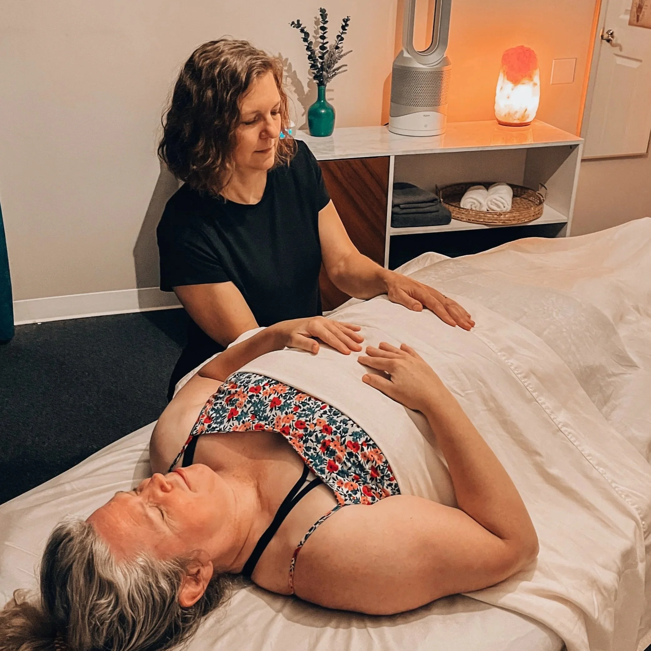 An elderly woman with gray hair lying on the floor with a younger woman, possibly a massage therapist, giving her a massage. The setting appears to be a relaxing room with decorative items, a diffuser, a salt lamp, and towels on a shelf.