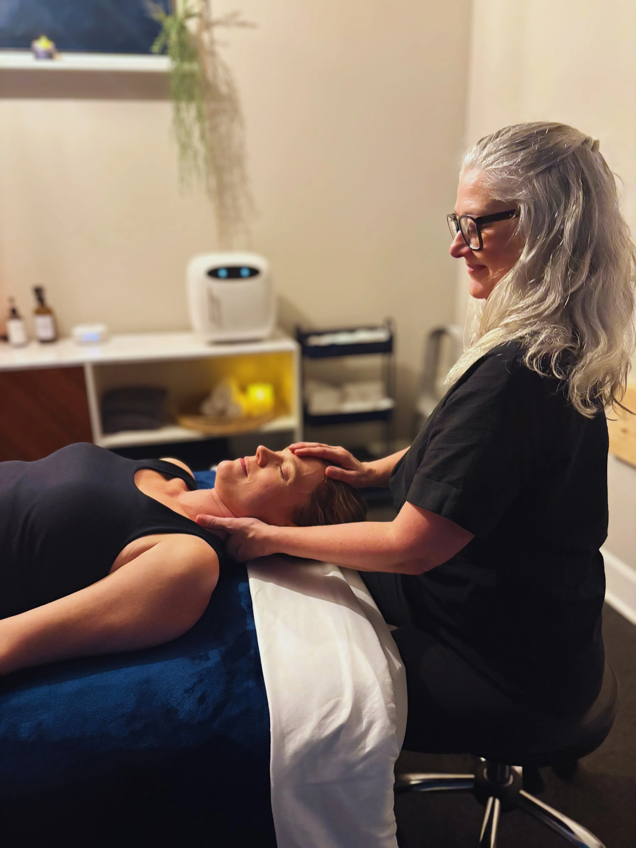 A woman with gray hair and glasses is giving a facial massage to another woman lying on a massage table in a spa or skincare clinic, with skincare products and a window in the background.