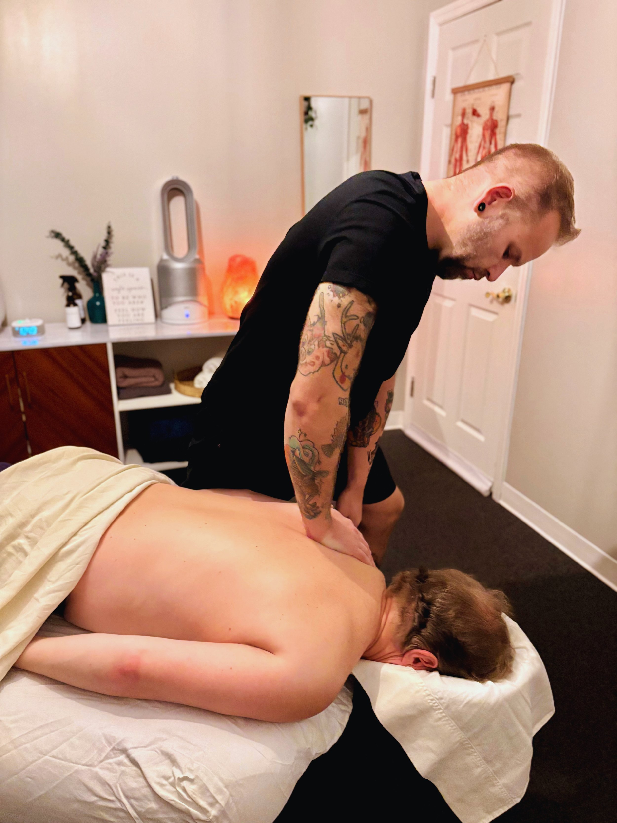A woman lying face down on a bed with her head turned to the side, receiving a massage from a man standing beside her. The scene takes place in a cozy room with soft lighting, a nightstand, and a decorated wall.