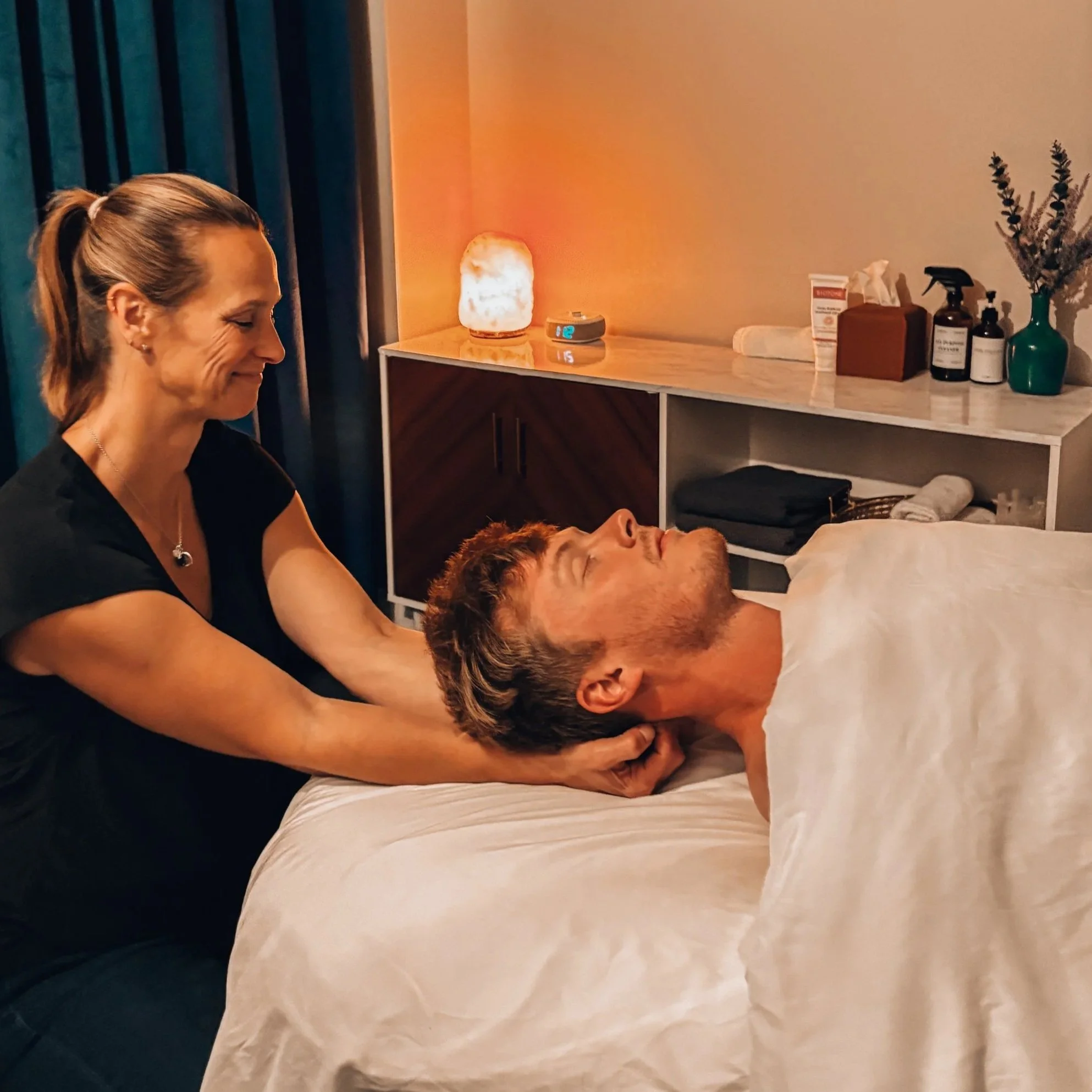 A woman providing a massage to a man lying on a massage table in a cozy room with warm lighting and decorative items on a shelf.