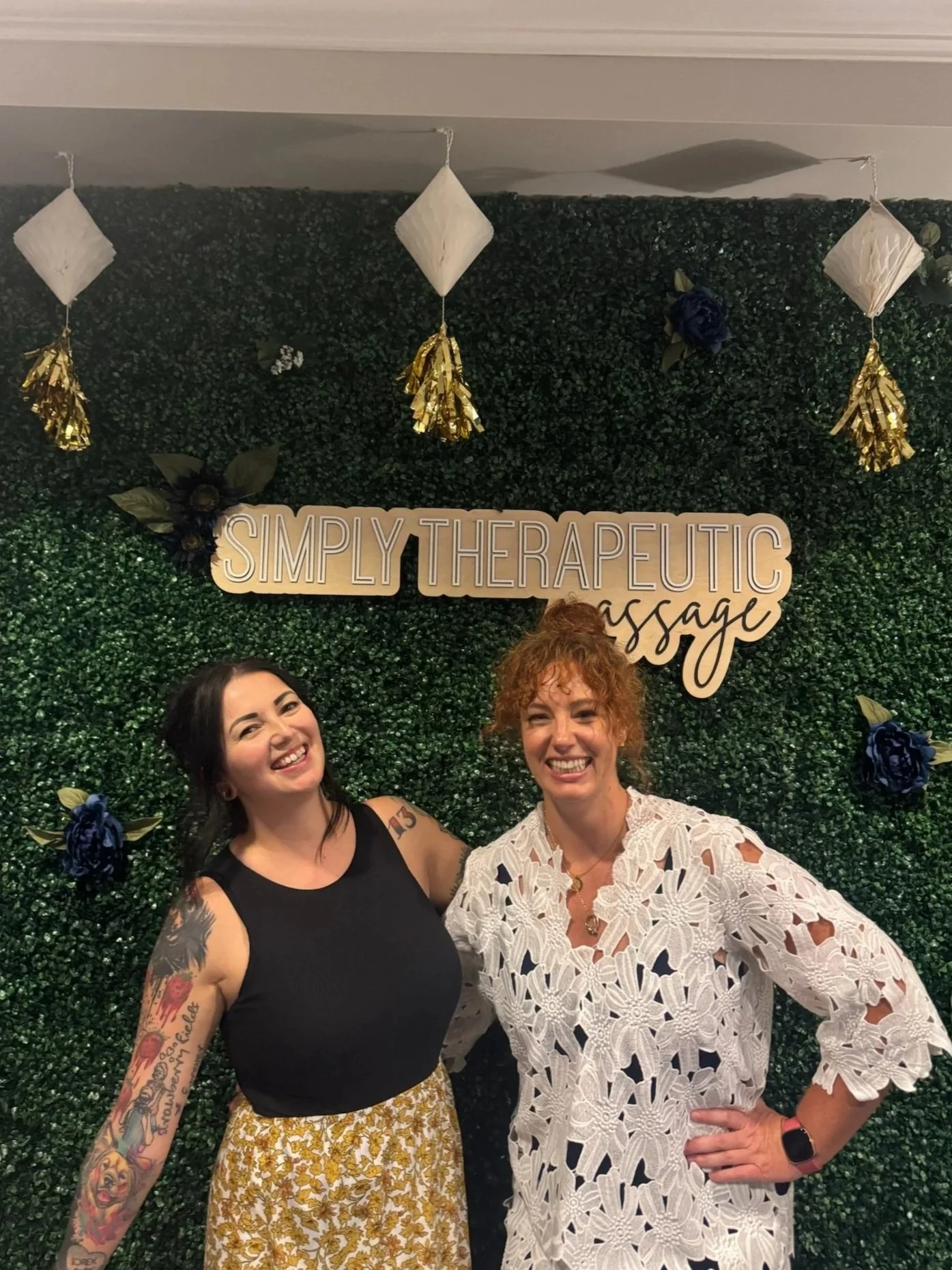 Two women smiling and standing close together in front of a green leafy wall with a sign that reads "Simply Therapeutic Massage" and decorative gold and dark blue flowers and paper lanterns hanging above.