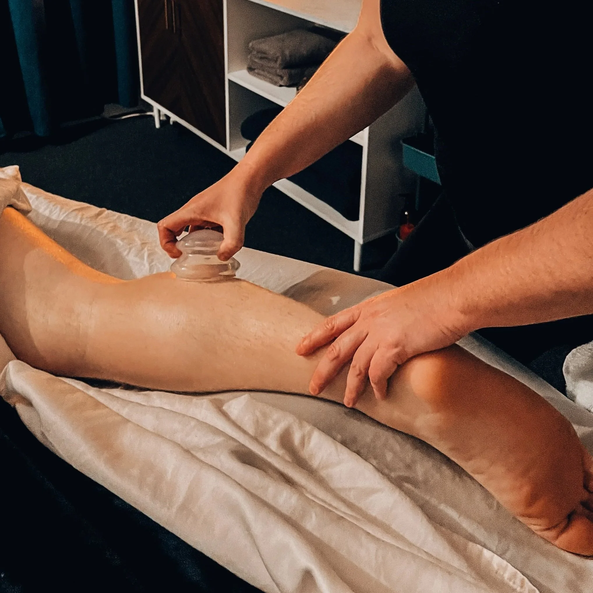 A person receiving a massage, lying face down on a massage table, with a massage therapist using a gua sha tool on their back. The setting appears to be a massage or wellness room with a cabinet and folded towels in the background.