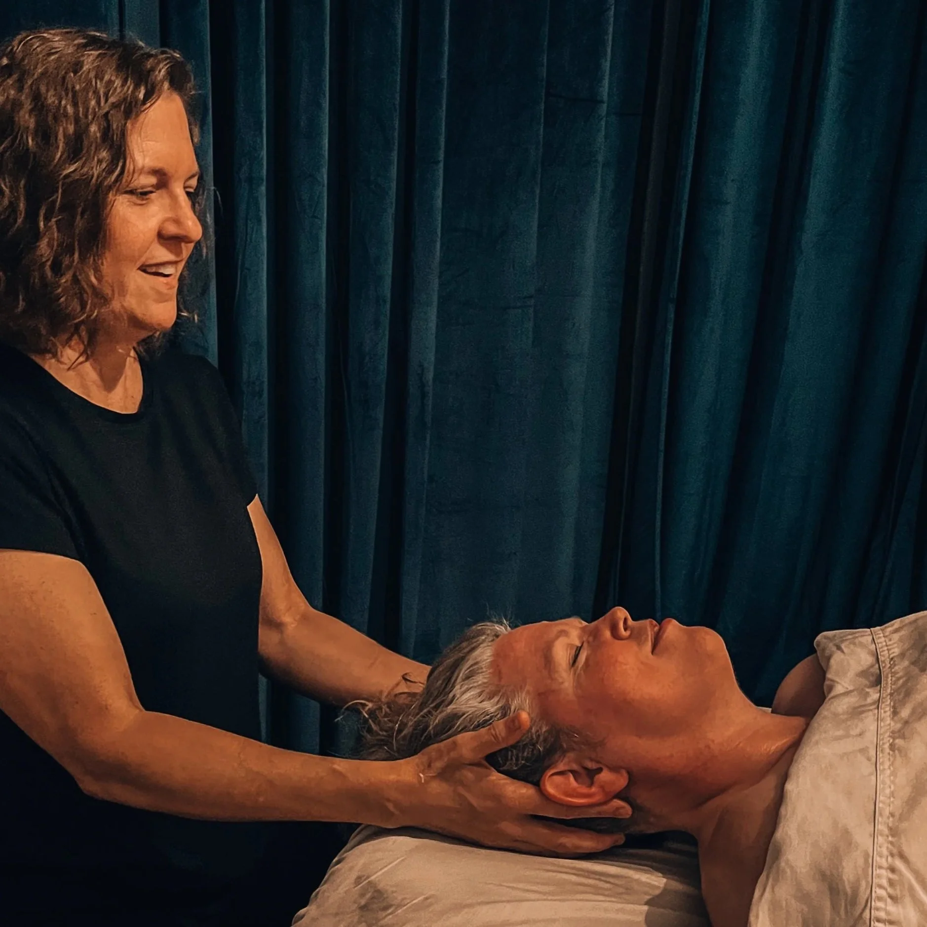 A woman giving a massage to an older man lying on a massage table with a blue velvet curtain in the background.