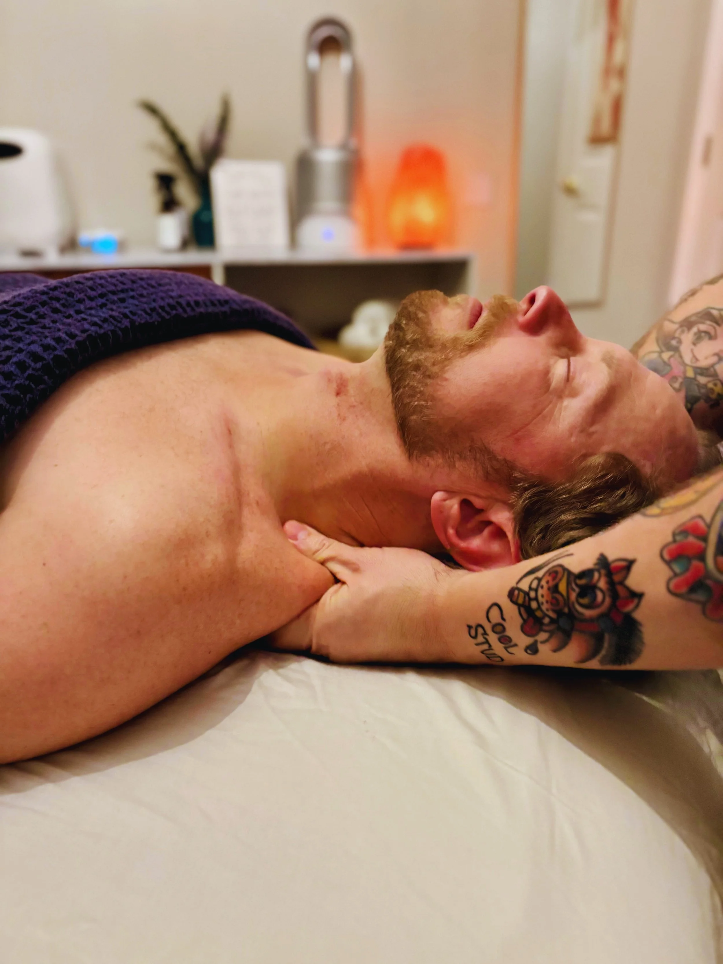 A person with tattoos lying on their side in a room, with their eyes closed, and a background that includes a countertop, a soap dispenser, and various items.