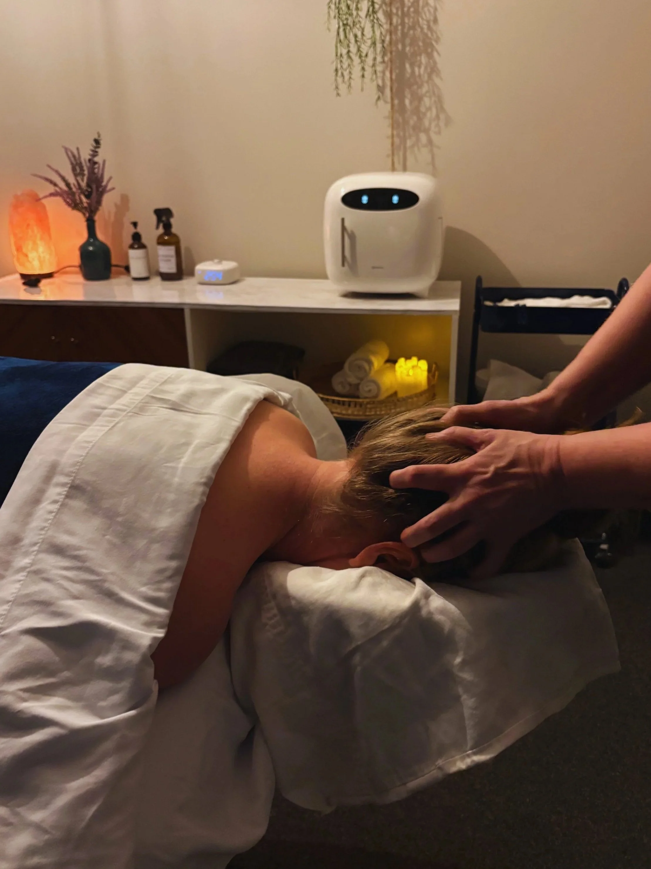 Person lying face down on massage table, receiving a head massage from a therapist in a wellness room. The room has warm lighting, decorative plants, a salt lamp, and wellness products on a sideboard.