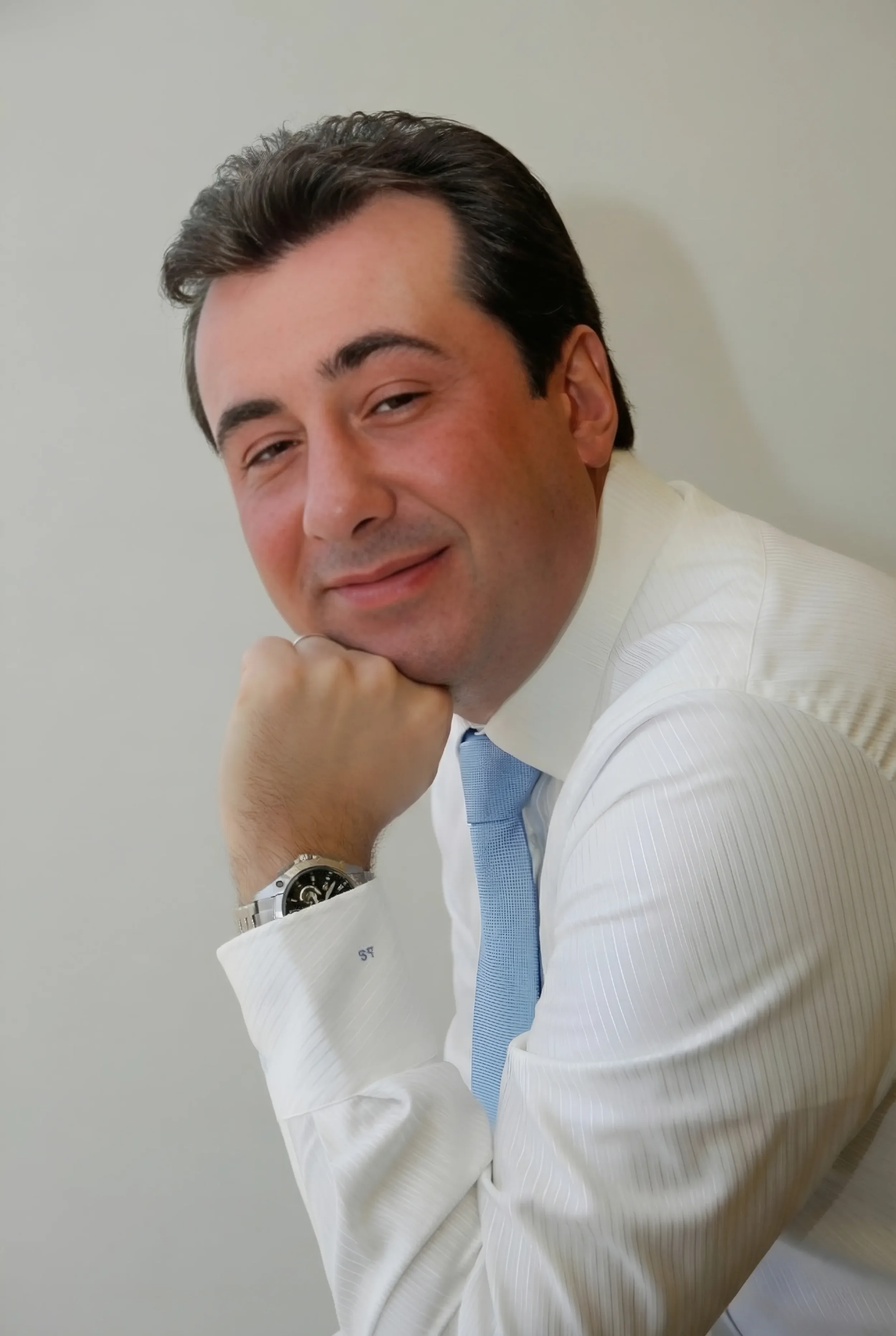 Andrea Stendardo | Senior full-stack engineer | Andrea Stendardo che sorride con la testa appoggiata sulla mano, indossa una camicia bianca e cravatta azzurra, con un orologio al polso.