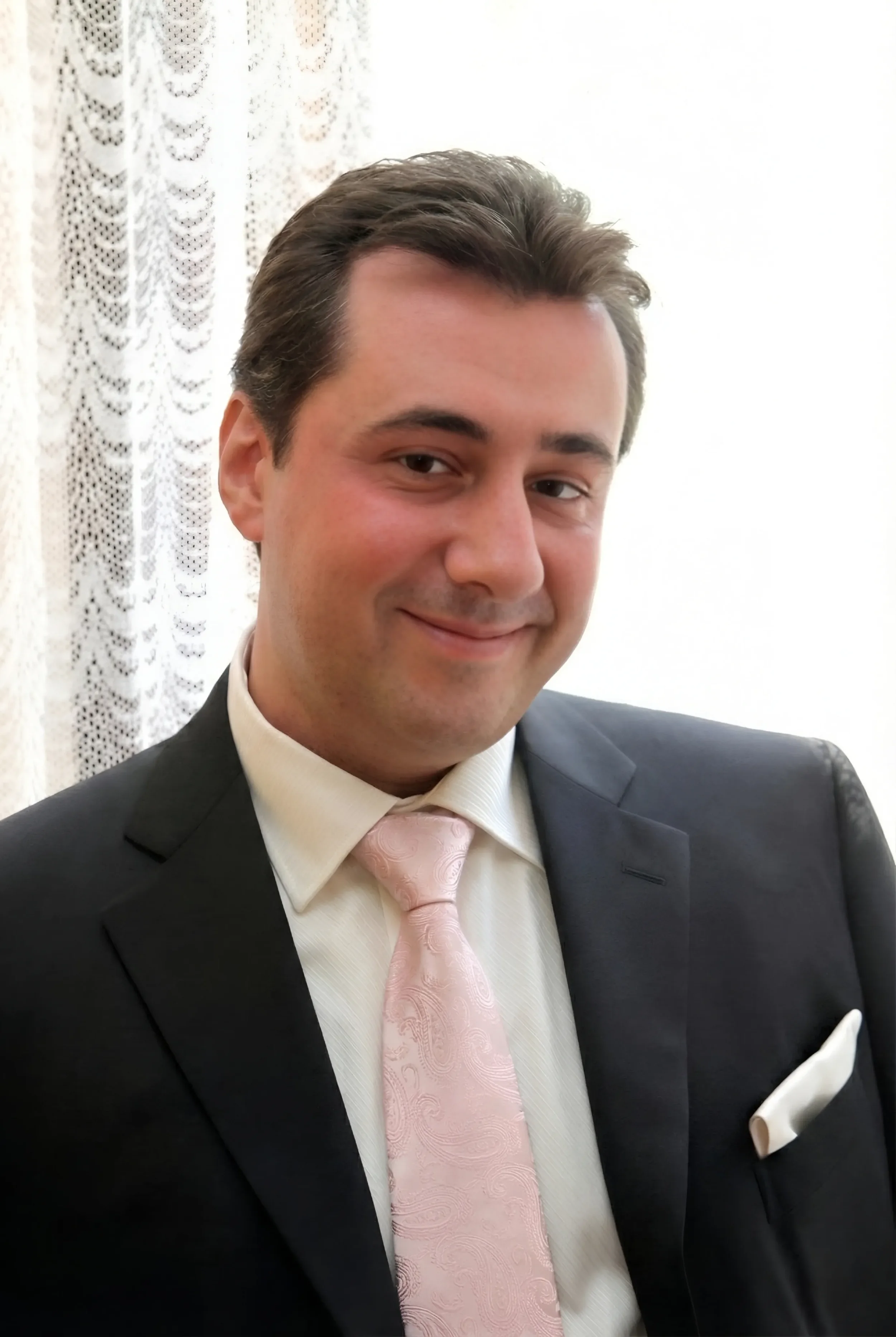 Andrea Stendardo | Senior full-stack engineer | Andrea Stendardo sorridente in abito formale con camicia bianca, cravatta rosa, in un ambiente con tende di pizzo bianco sullo sfondo.