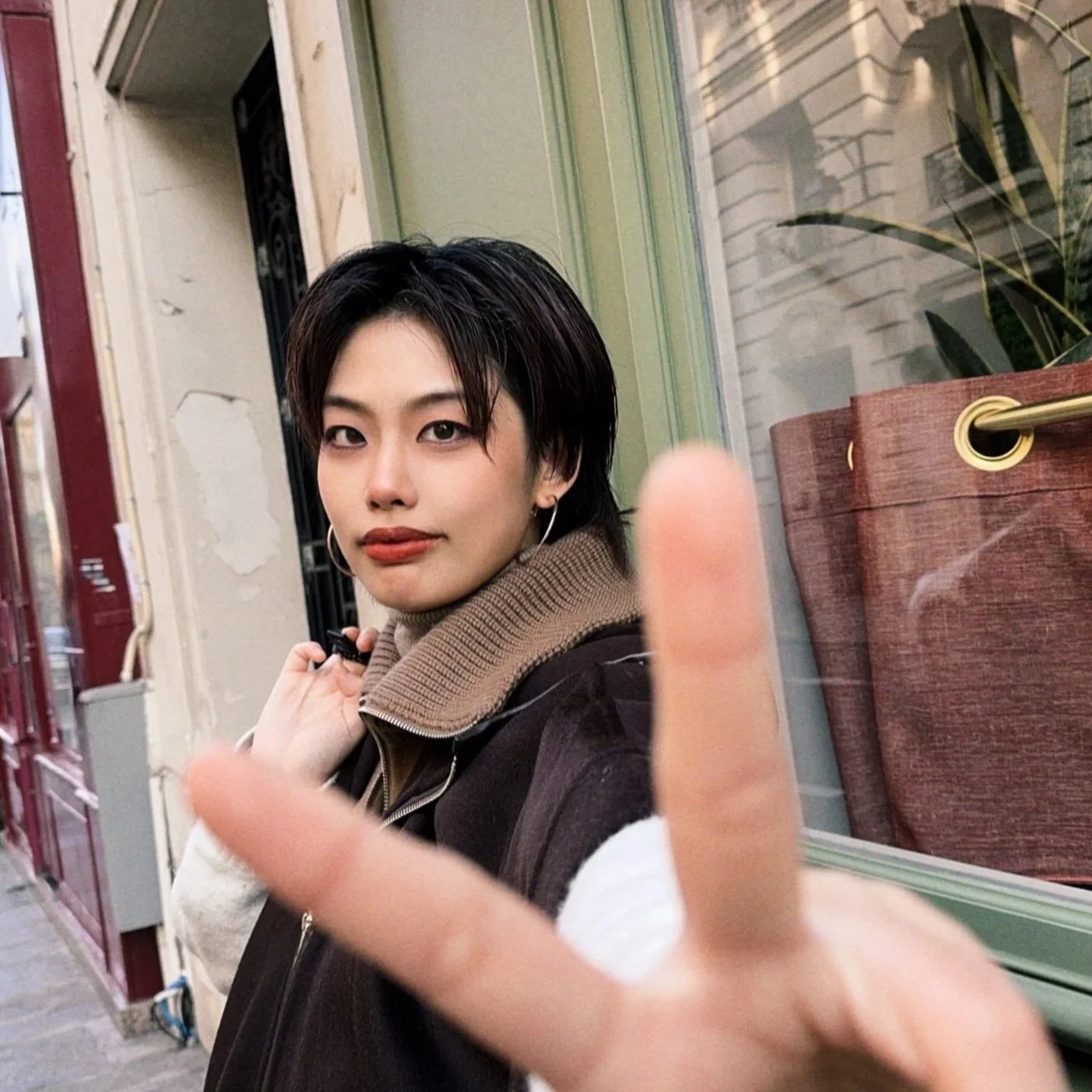 A young woman with short black hair and earrings making a peace sign with her hand in front of a window, sitting on a sidewalk with buildings in the background.
