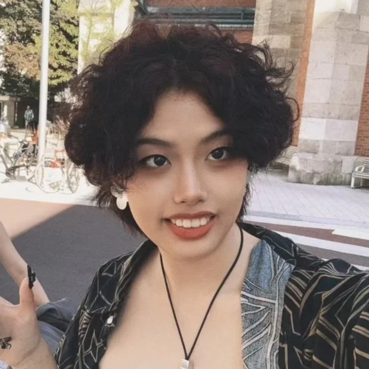 Young woman with short curly dark hair, smiling, wearing white earrings, black choker with a pendant, patterned black shirt, outdoors near brick building and bicycles.