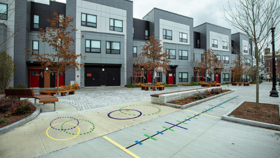 Outdoor urban scene with modern apartment buildings, leafless trees, benches, and children's play area with hula hoops and hopscotch on paved ground.