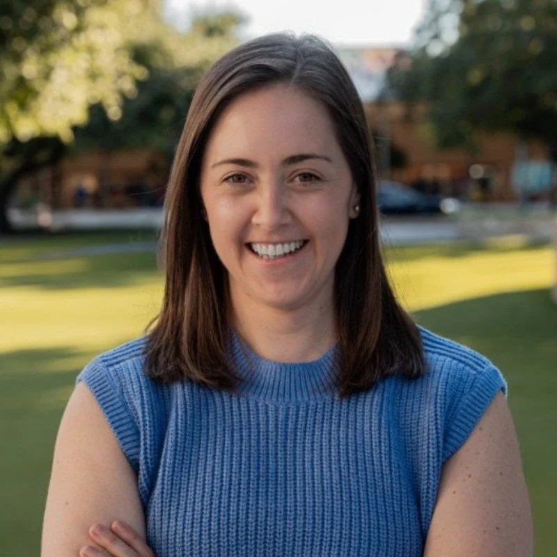 Smiling woman with shoulder-length brown hair wearing a sleeveless blue knit top, outdoors with trees and a grassy area in the background.