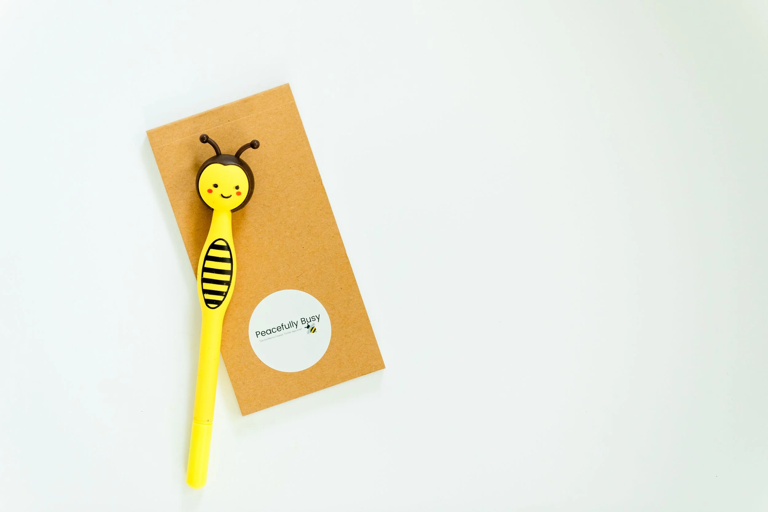 Yellow pen with a cute bee face at the top and black striped patterns on the body, placed on a brown paper envelope with a label that reads "peacefully Busy" and a small bee illustration, on a white background.