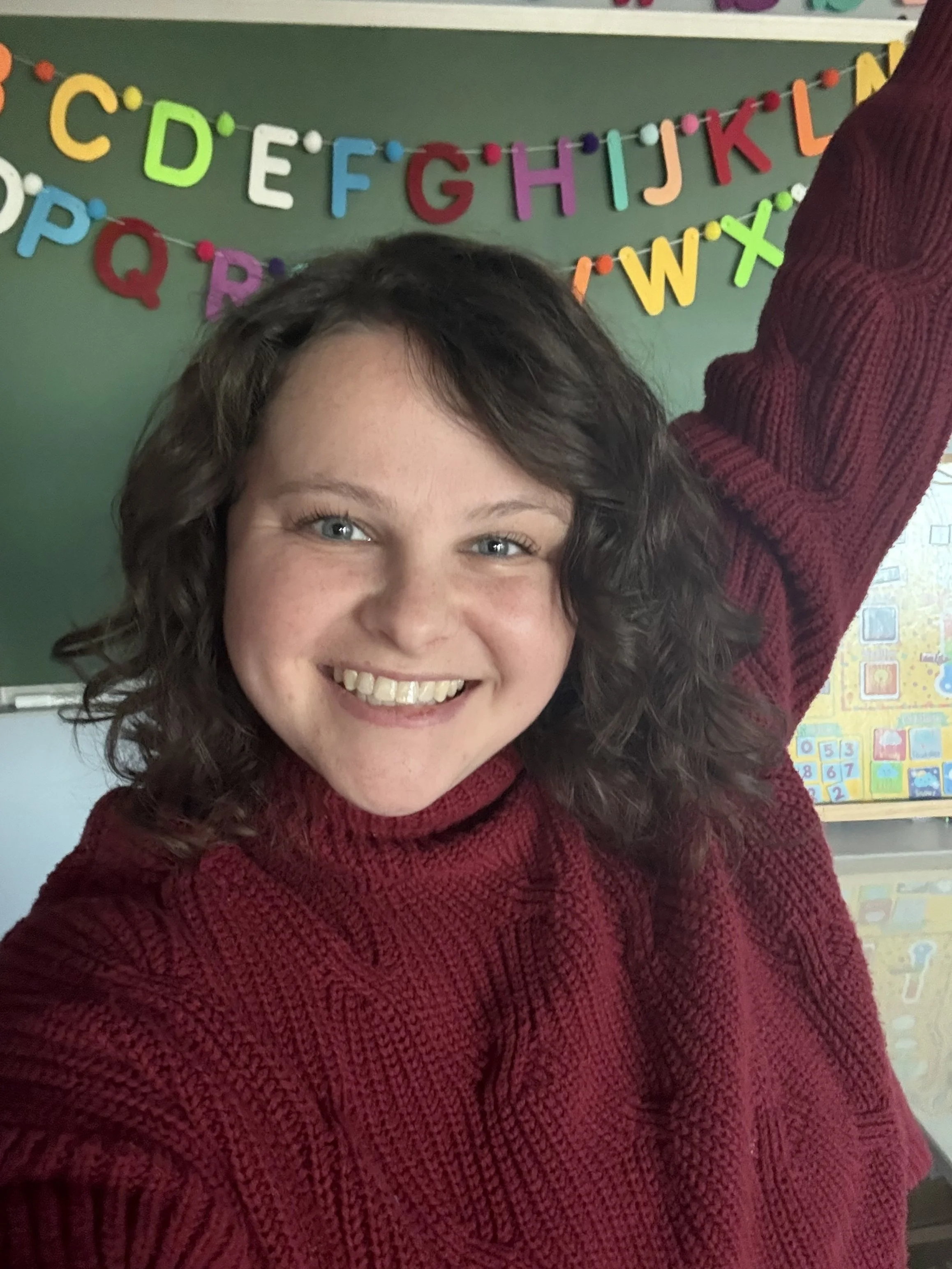 A smiling woman in a red sweater with curly hair, raising her arm, in a classroom with colorful alphabet decorations on the chalkboard behind her.