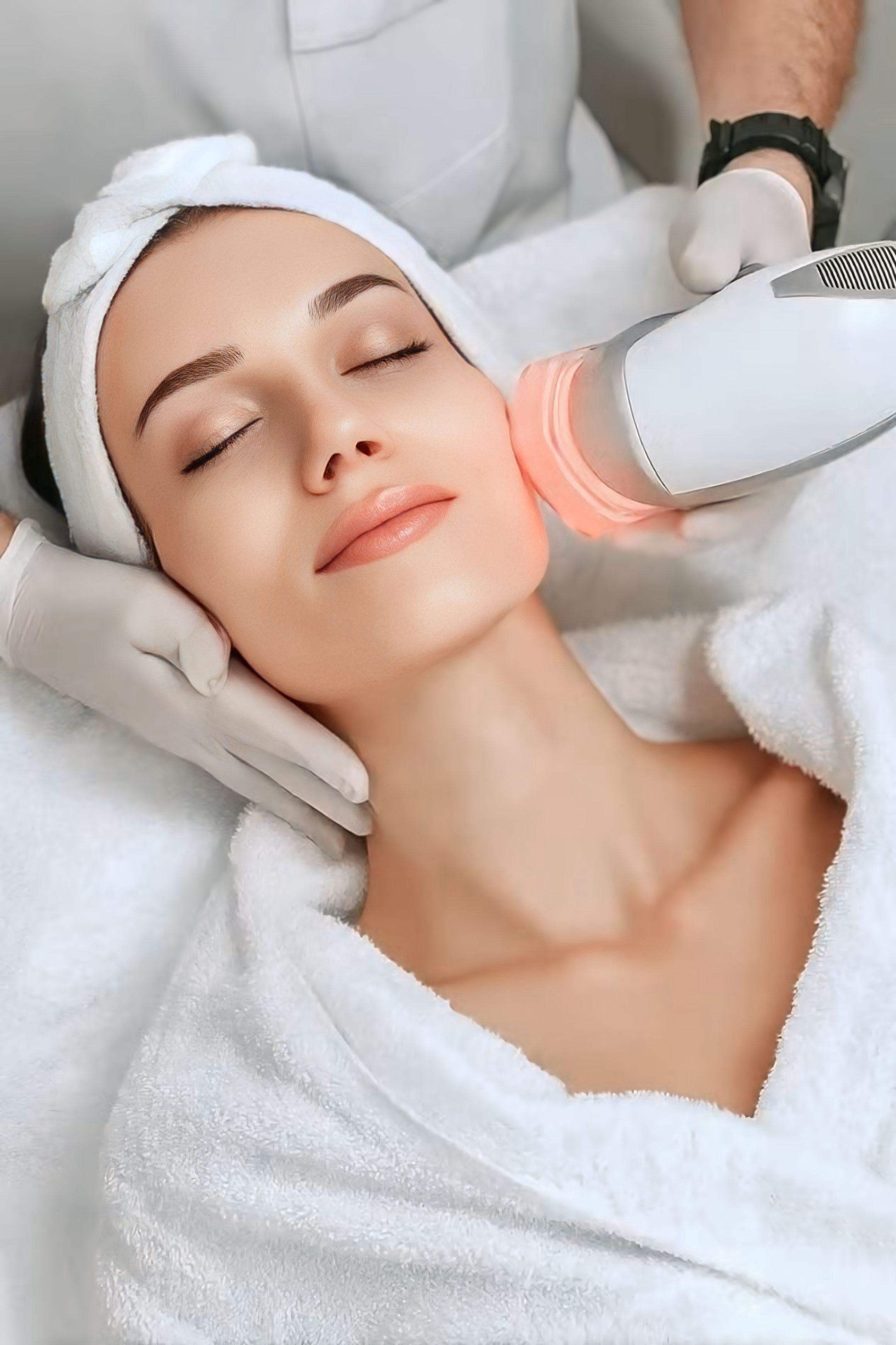 Mujer en tratamiento facial de estética, relajada con ojos cerrados, recibiendo tratamiento con luz LED en la cara en un entorno clínico.