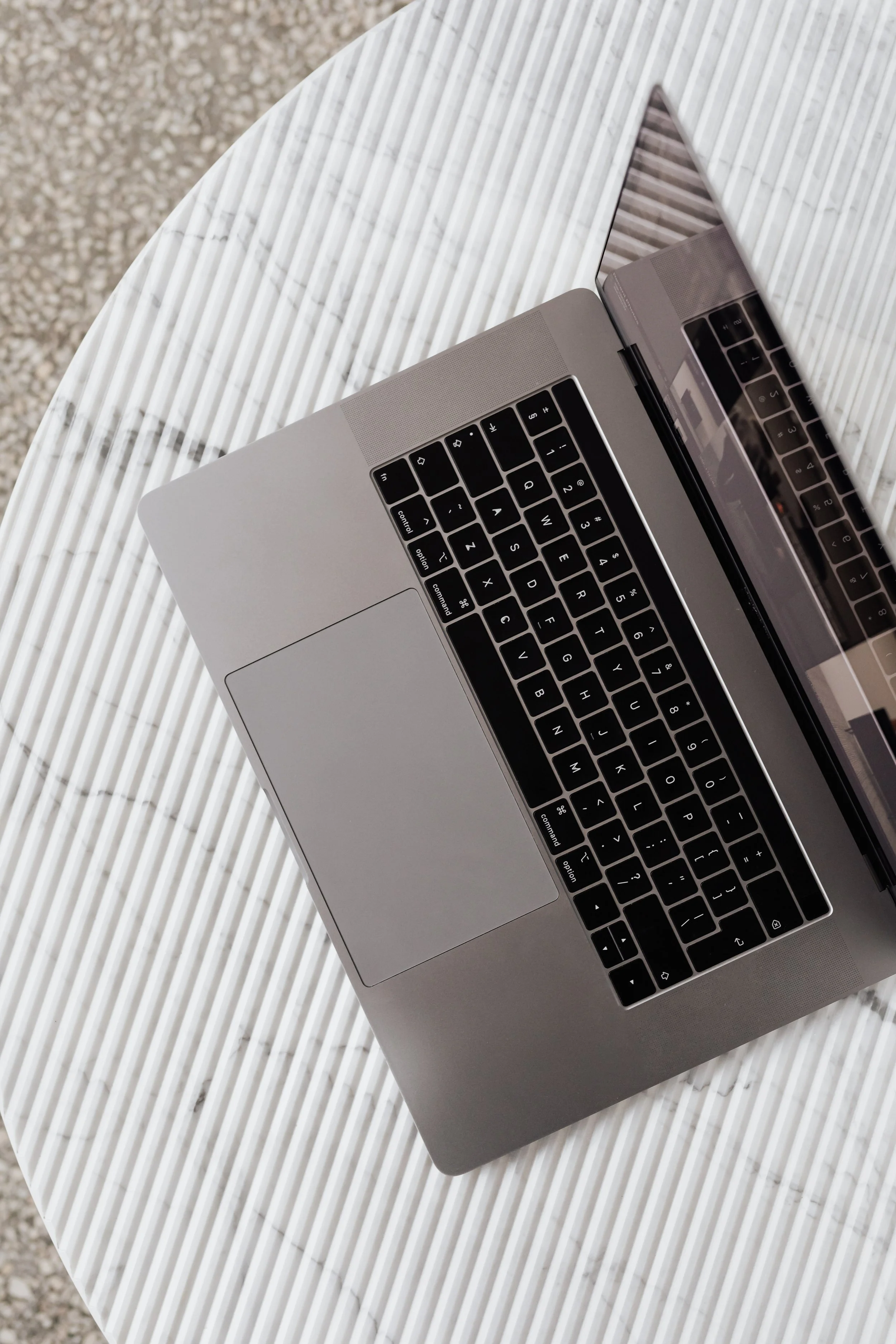 A silver laptop with black keyboard on a striped white and light gray table, showing a reflection of the keyboard on the screen.