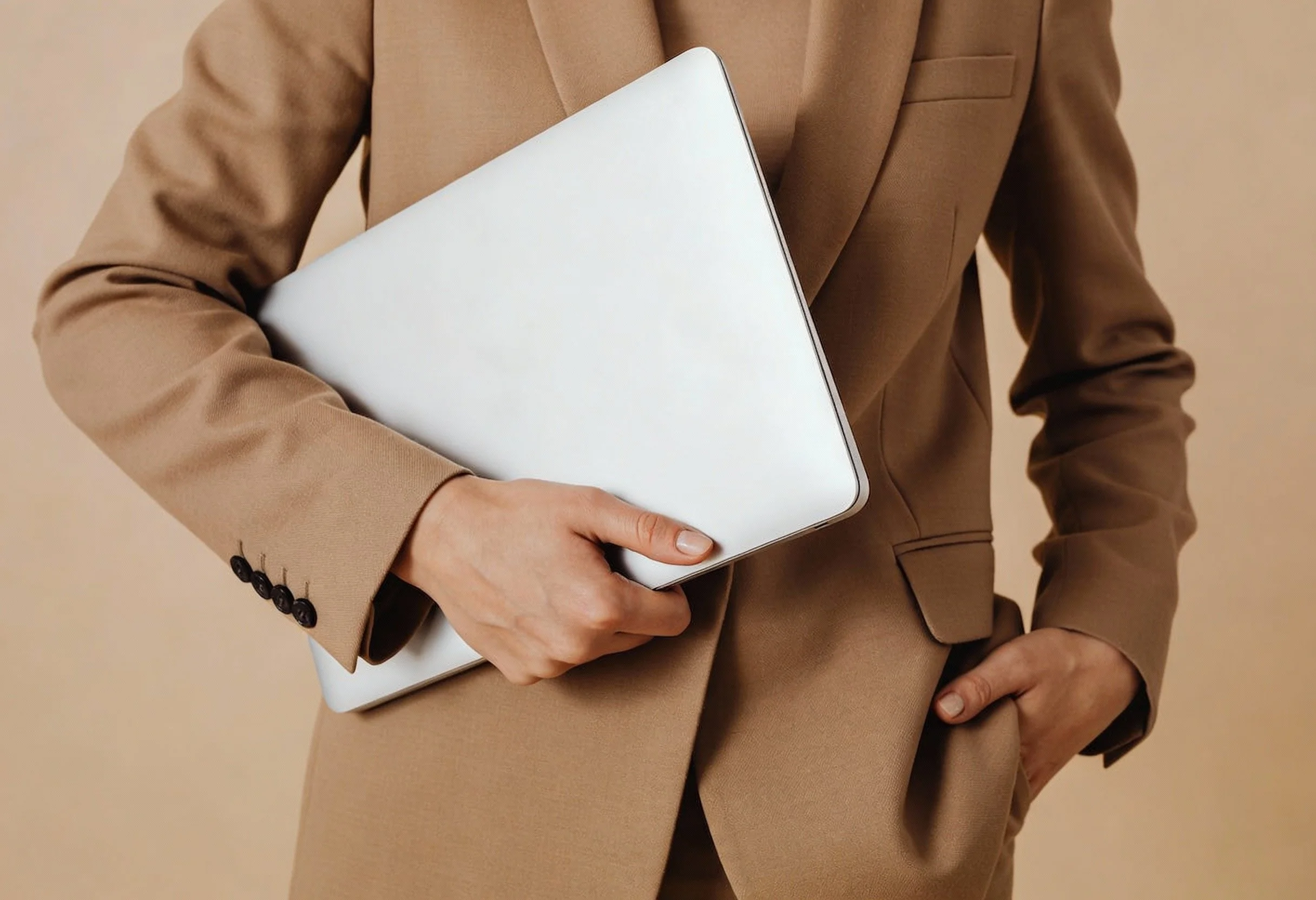 A person dressed in a beige business suit holding a closed silver laptop in their right hand, with their left hand in their pocket.