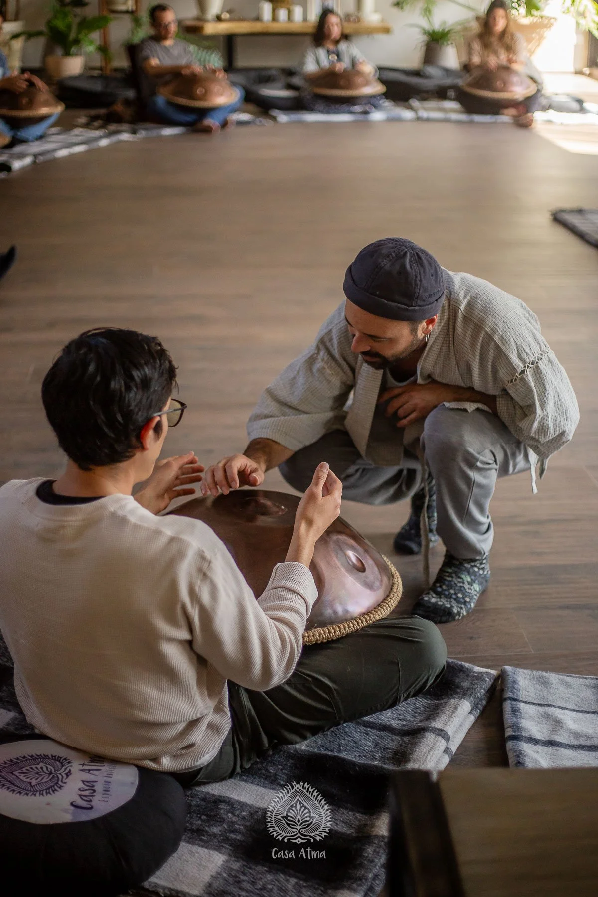 Dos personas en el suelo tocando un hang drum, con un grupo de personas en el fondo en actitud similar en una sala de madera y plantas.