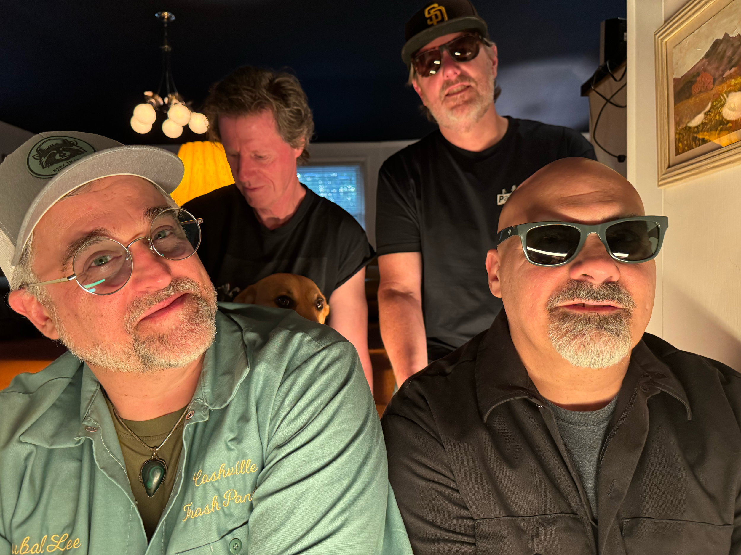A group of five men with a dog, posing for a photo in a dimly lit room. They are wearing sunglasses and casual attire, with a dark ceiling and warm lighting in the background.
