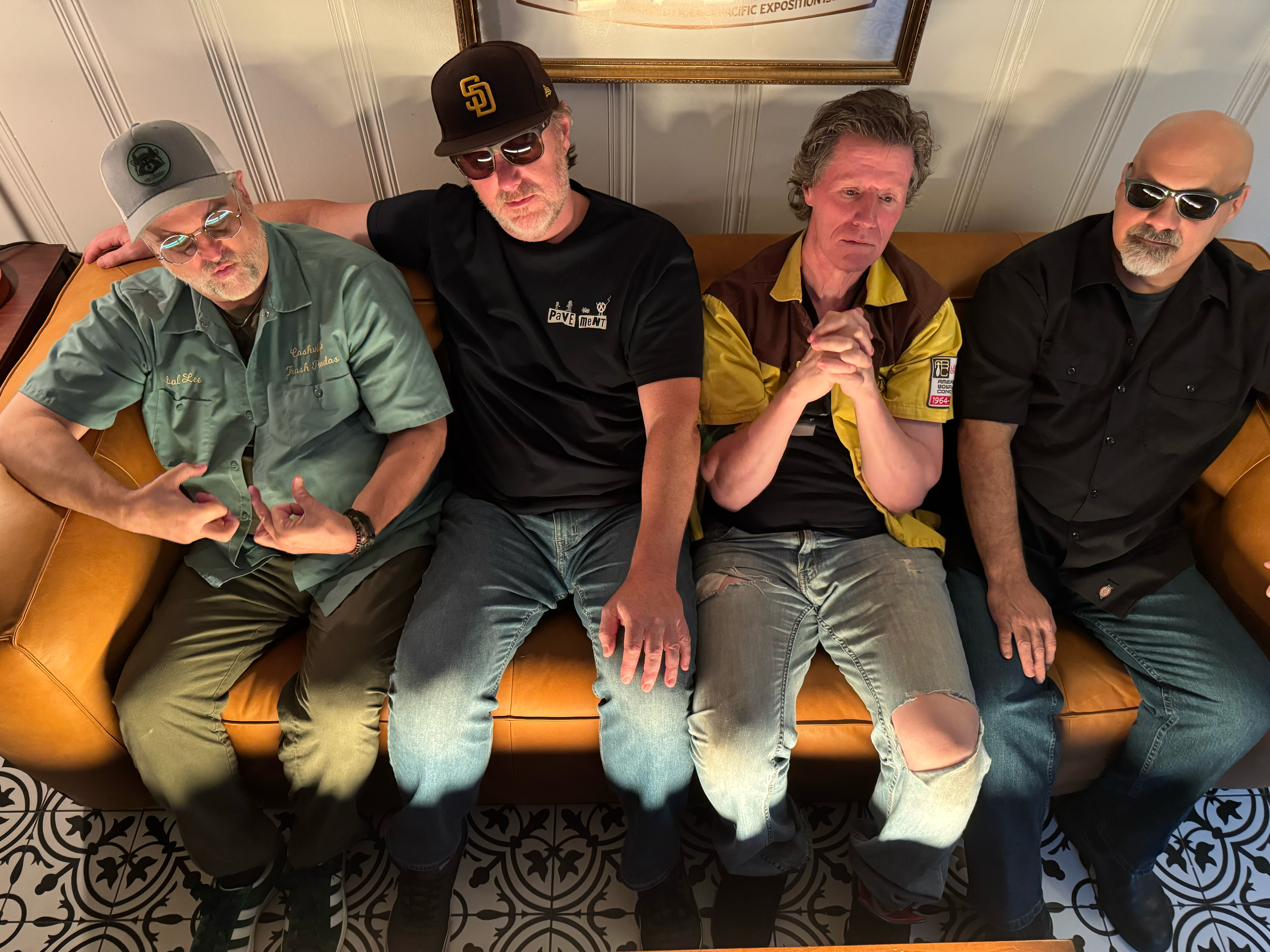 Four men sitting on a tan leather couch, making various hand gestures, in a room with white paneled walls and black and white patterned floor tiles.
