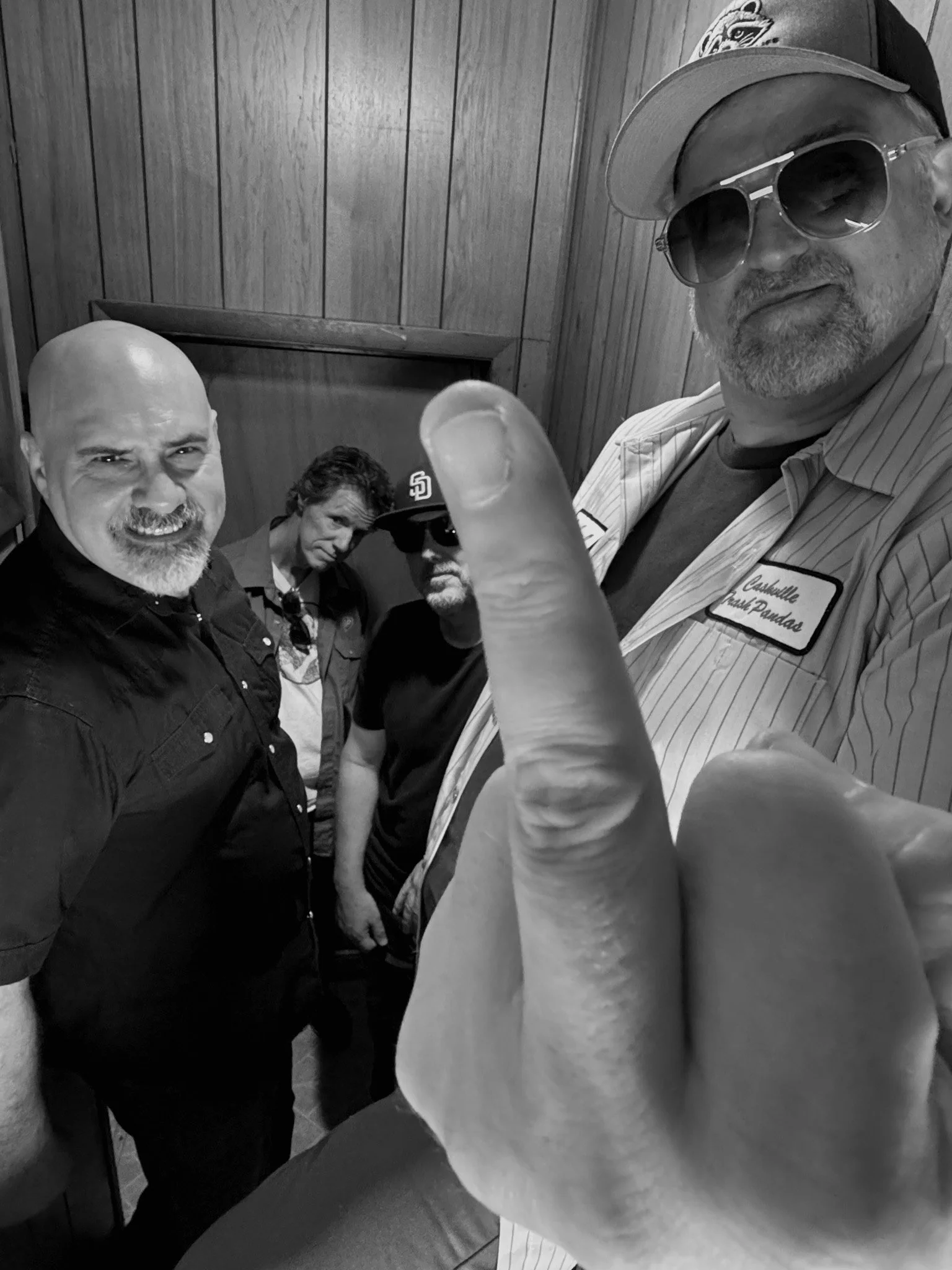 Four men sitting together indoors, with a wooden wall in the background. One man in the foreground is taking a selfie, wearing sunglasses and a cap, with a name tag that reads 'Ashtonville' and 'Jesse Dandas'. The other men are looking at the camera, with one wearing sunglasses and a cap, and the others have various hairstyles and facial hair.