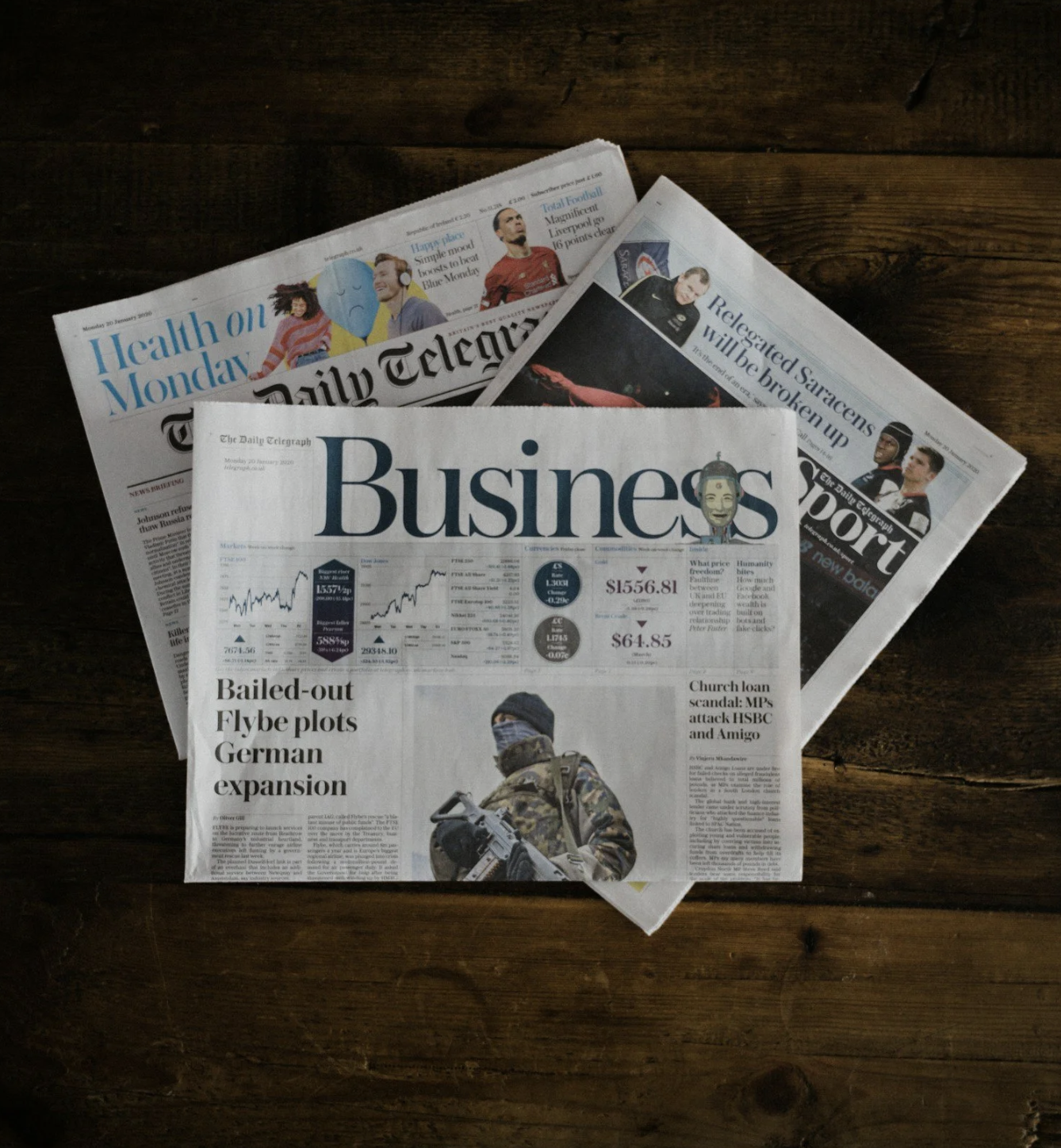 Four newspapers stacked on a dark wooden table, with the top one titled 'Business' featuring stock market reports and a photo of a soldier in military gear.
