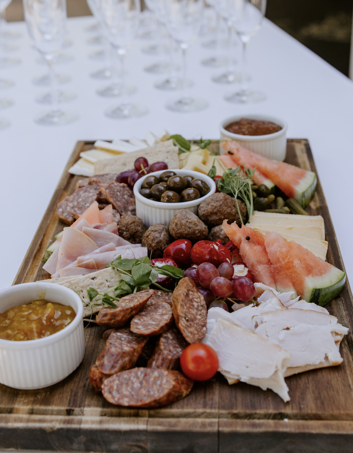 Charcuterie board with cheeses, sliced meats, grapes, watermelon, olives, cherry tomatoes, and dips on a wooden platter.
