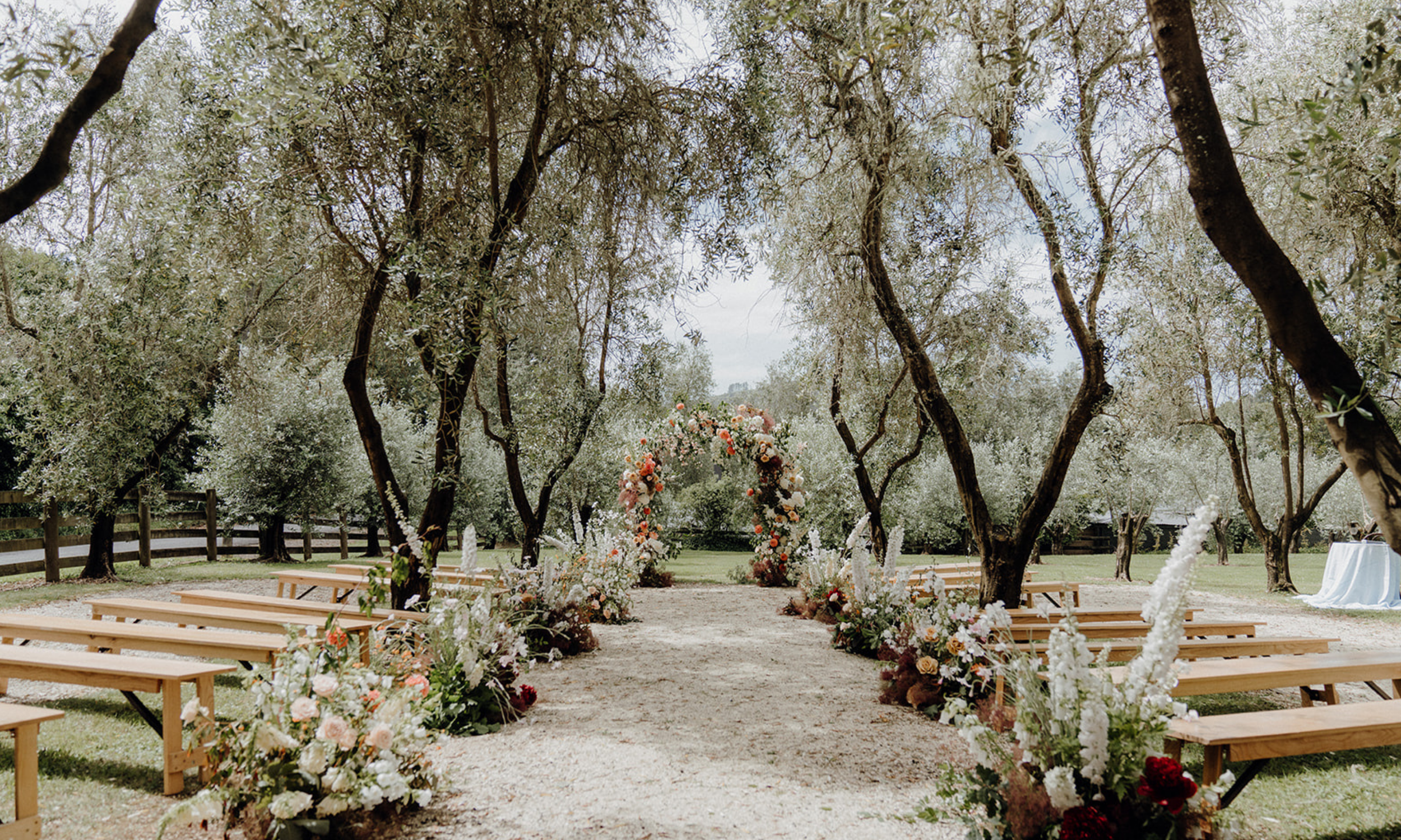 Outdoor wedding setup with an aisle decorated with floral arrangements leading to a floral arch, surrounded by trees and benches.