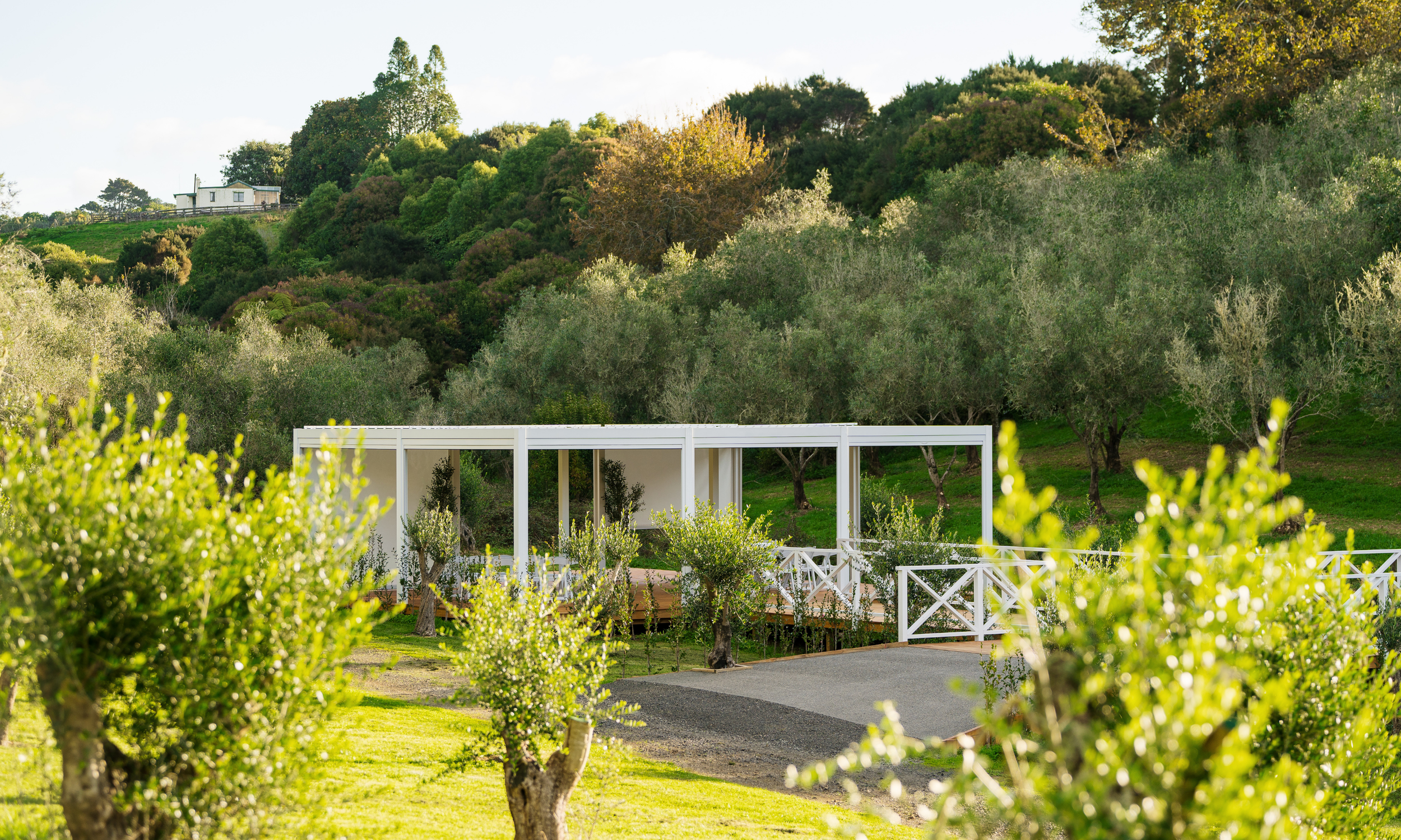 A modern white outdoor patio structure in a lush green garden with trees and rolling hills in the background.