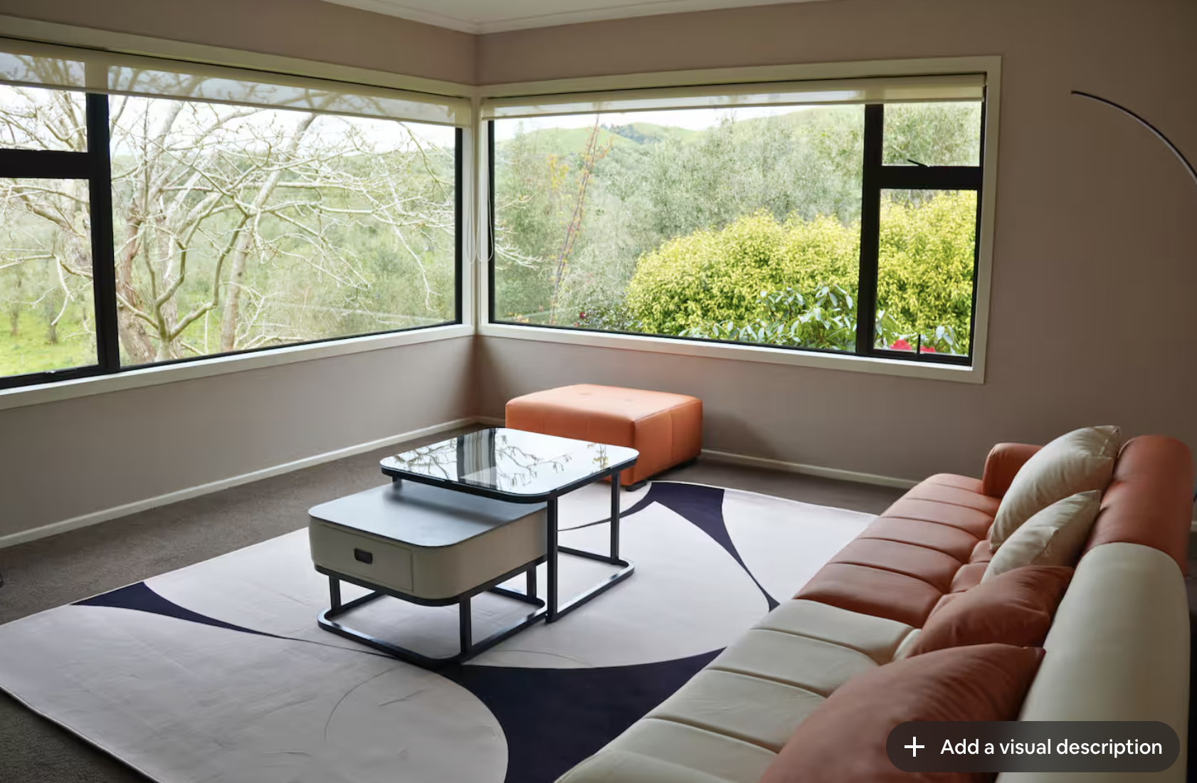 Living room with large corner windows showcasing a view of trees and green foliage outside, a white and orange sofa with beige pillows, a glass-topped coffee table with a black metal frame, an orange ottoman, and an abstract white and black rug.