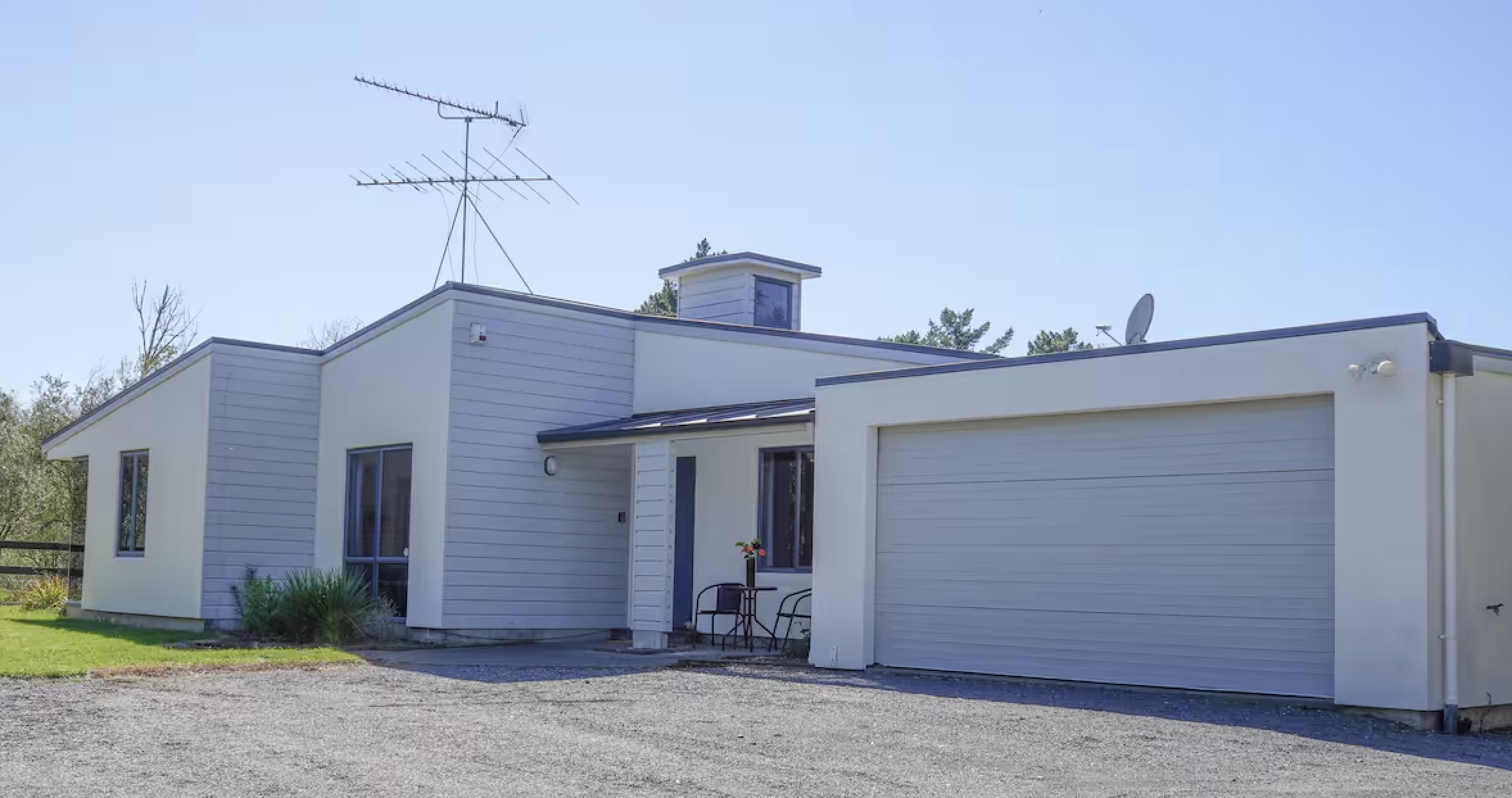 A modern detached house with a white exterior, gray panels, and a large garage door. There is a small outdoor seating area with two chairs and a table with flowers. The house has an antenna on the roof and a satellite dish on the side. Green grass and trees surround the house, and the sky is clear.