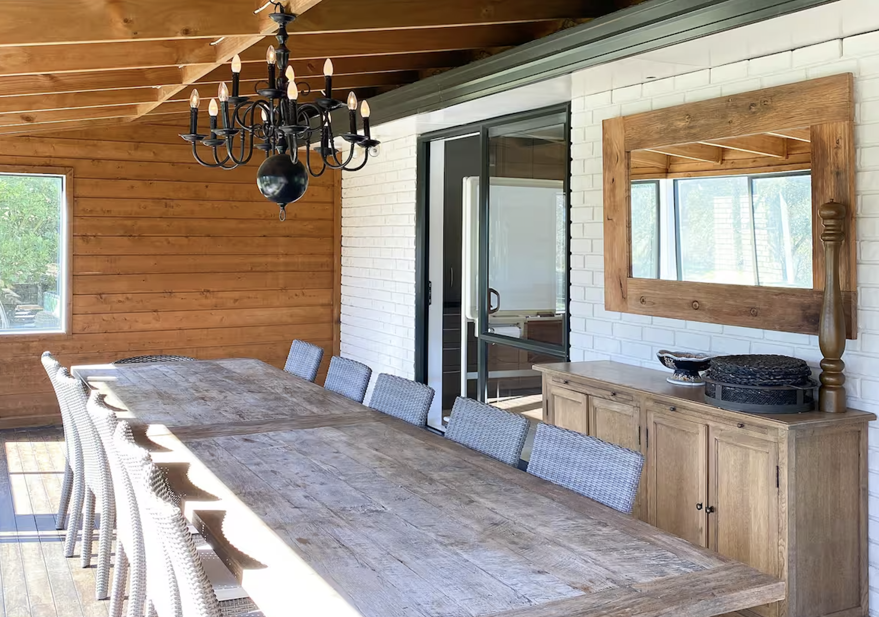 A dining room with a rustic wooden table, gray woven chairs, a black chandelier with candle-shaped lights, a wooden sideboard with a mirror, a white brick wall, and a sliding glass door leading outside. Sunlight is streaming in through the windows.