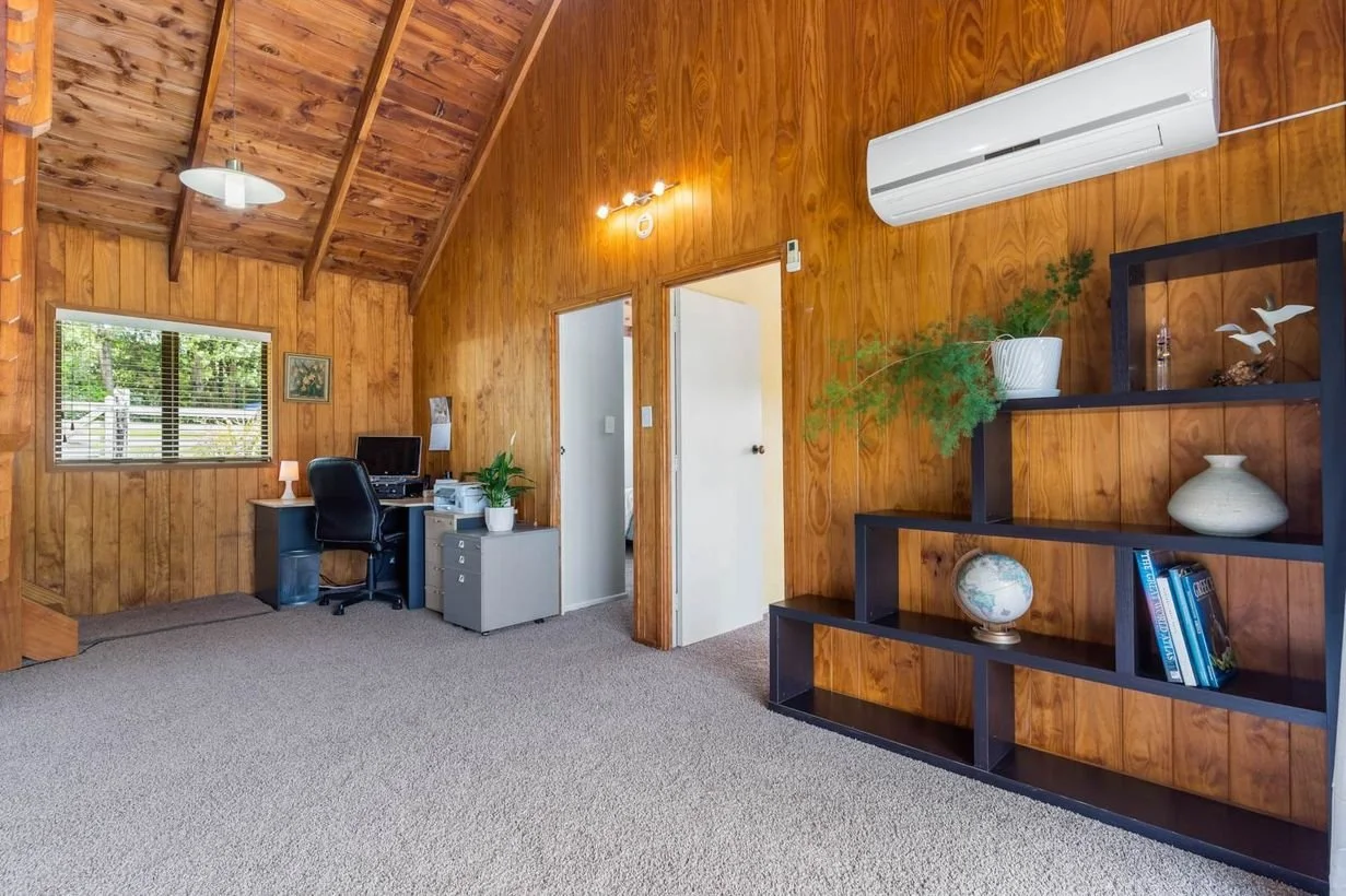 Wood-paneled room with desk, computer, and office chair near window, black shelving unit with decorative items and books, and a white air conditioning unit on the wall.